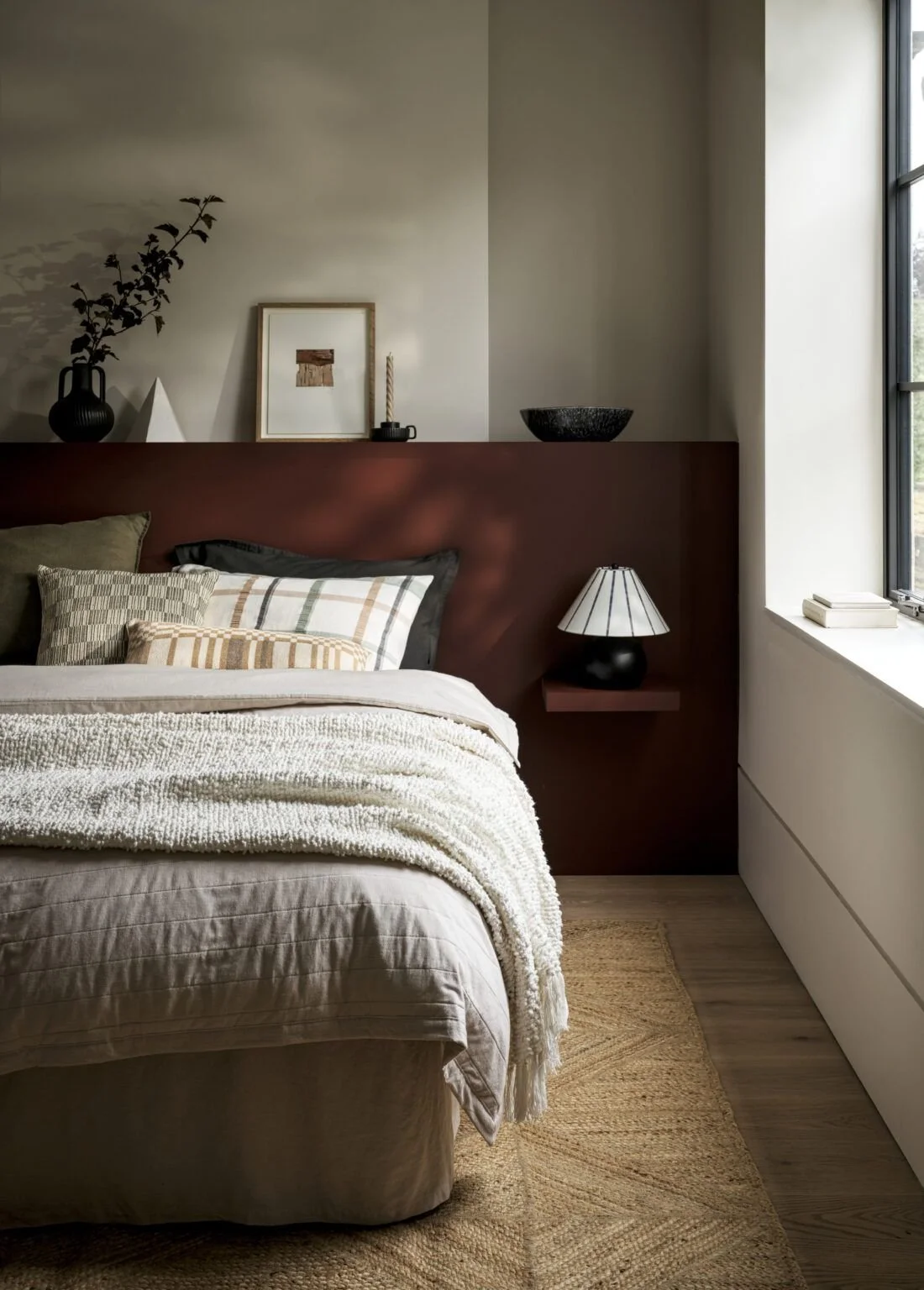 A cozy bedroom with a bed, decorative pillows, a textured blanket, a nightstand with a lamp, wall art, and a window with a ledge.