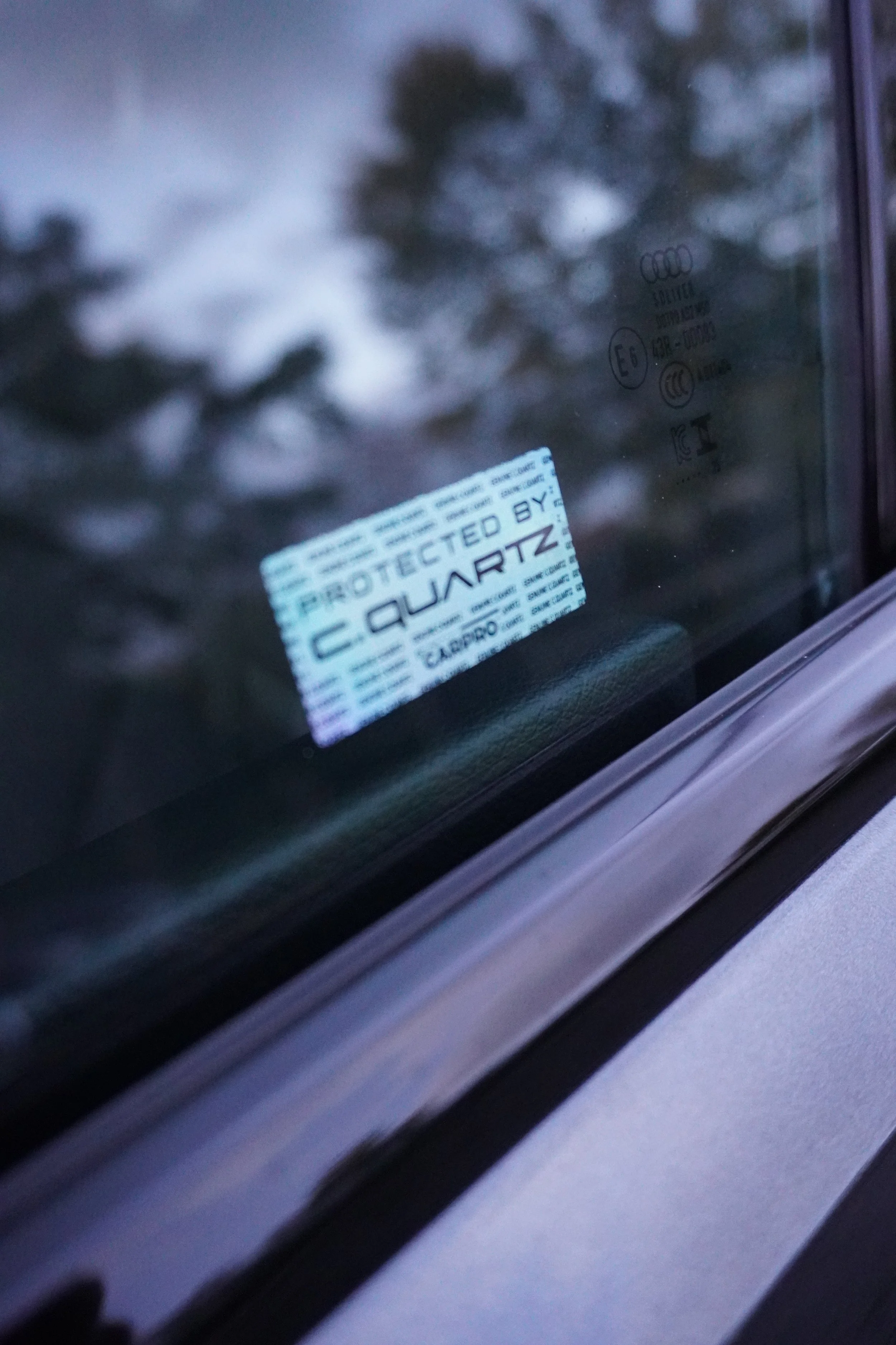 Close-up van een autoglas met etiketten, onder andere met de tekst 'PROTECTED BY' en 'C.QARTZ'.