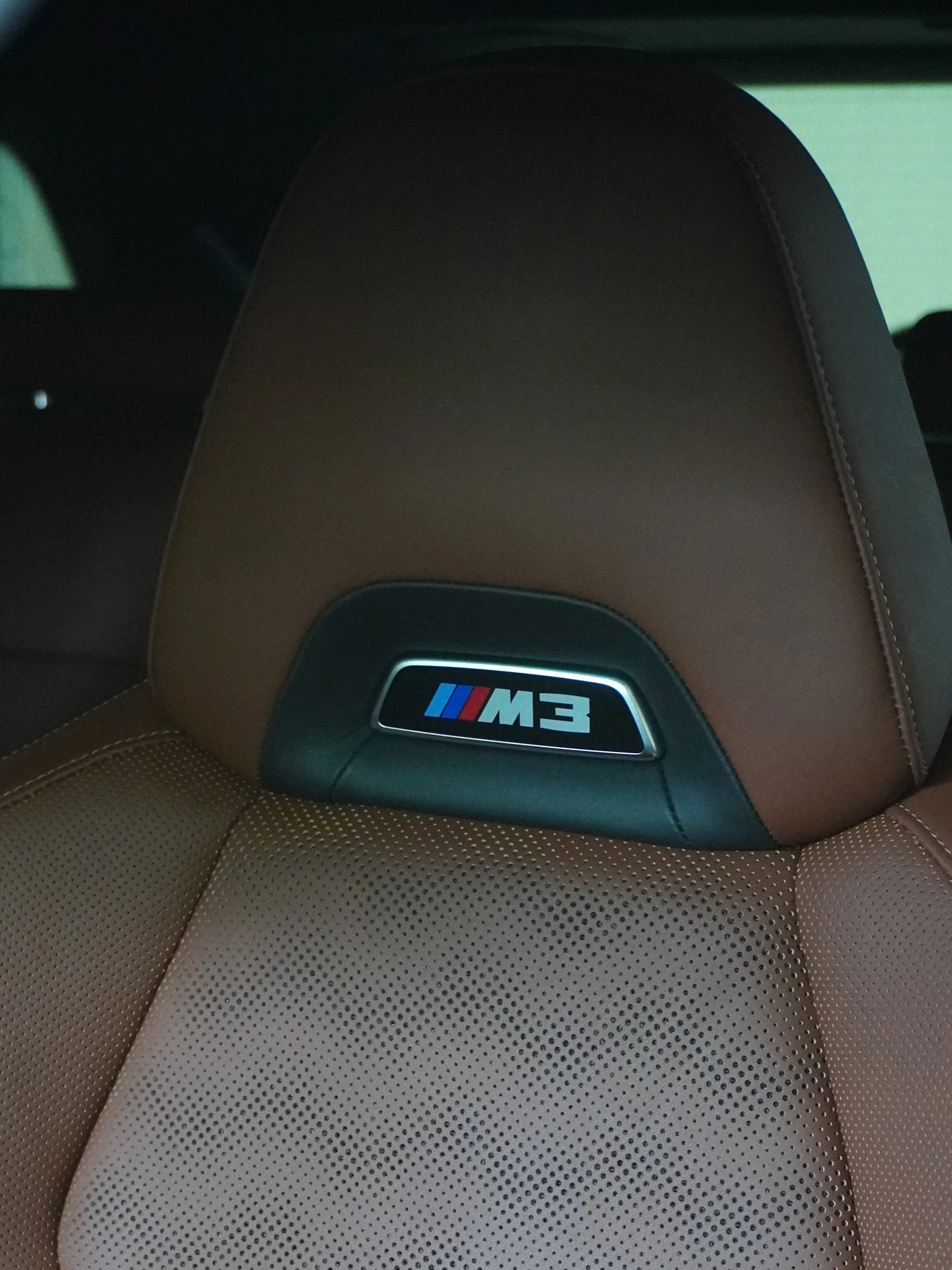 De foto toont het hoofdsteuntje van een sportwagen, waarschijnlijk een BMW M3, met een logo en gestikte details. De stoelen zijn bruin leer met perforaties.