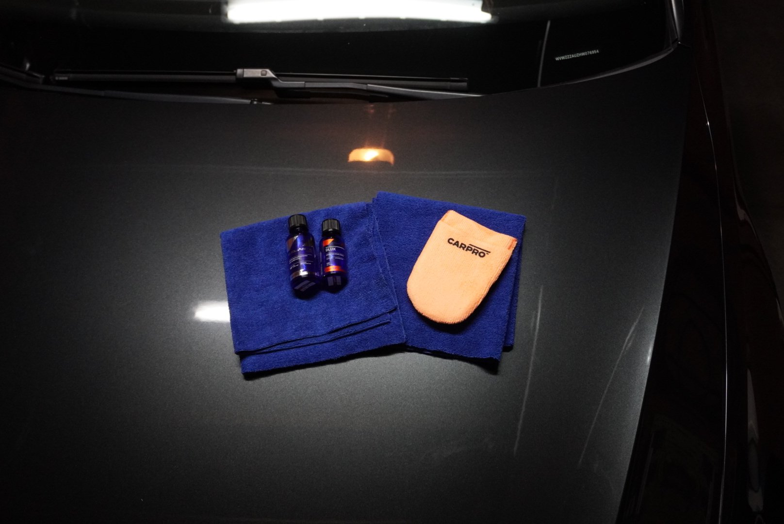 Twee kleine flesjes vloeistof, een blauwe handdoek, een oranje spons en een leren poetsdoek, allemaal op een donkerautokap.