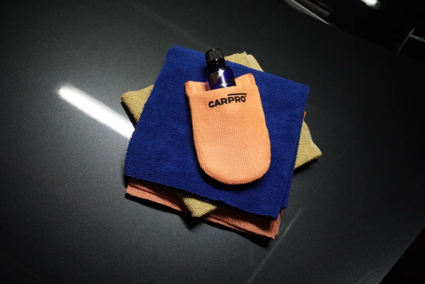Een flesje autoverzorgingsmiddel, gelegd op twee gevouwen microvezeldoeken, met een blauw en een gele doek, op een zwarte carrosserie.