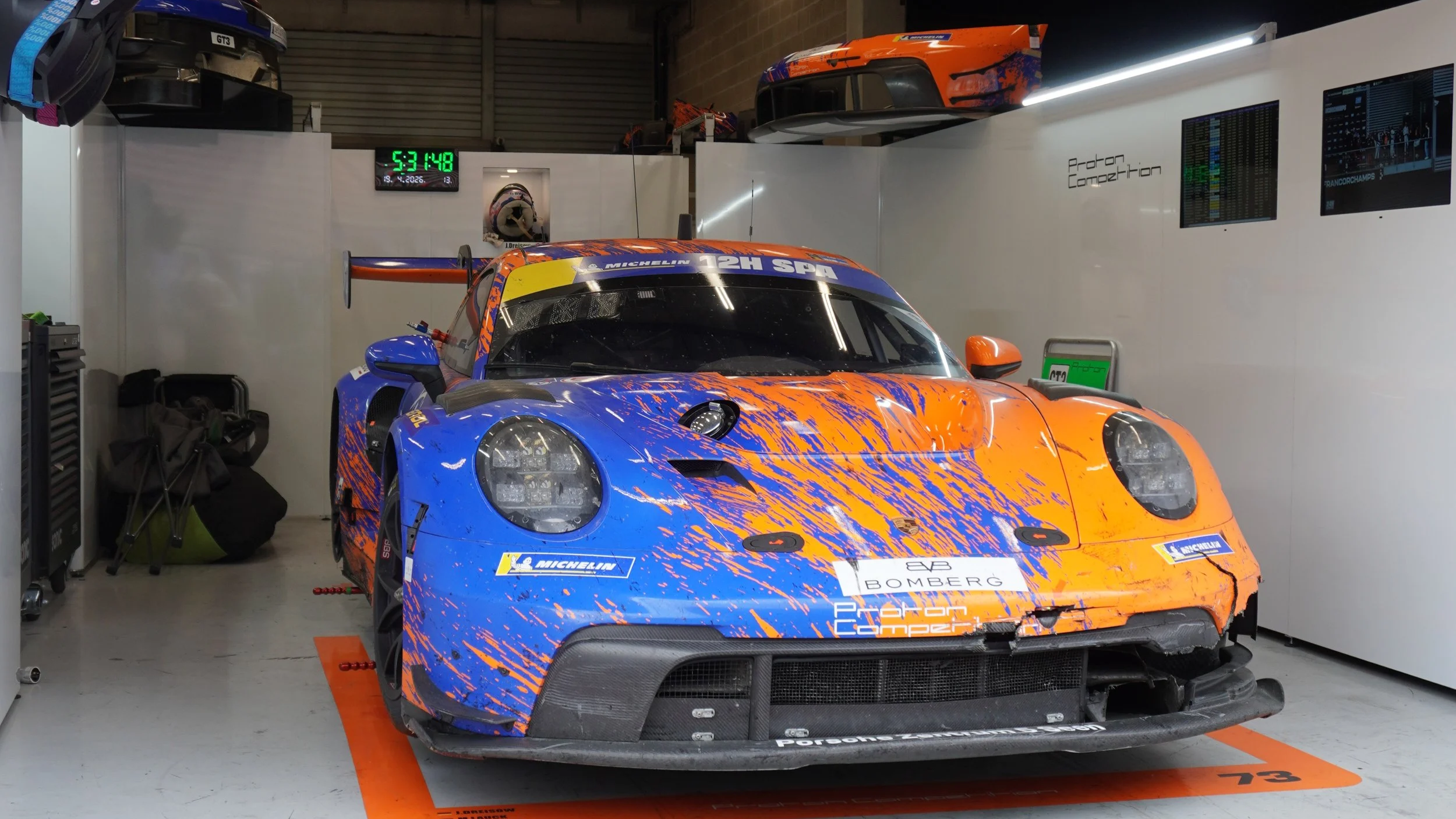 Raceauto in een pitbox met blauwe en oranje verfspatten, digitale klok en schermen met gegevens, en racemotorhelm op de muur.