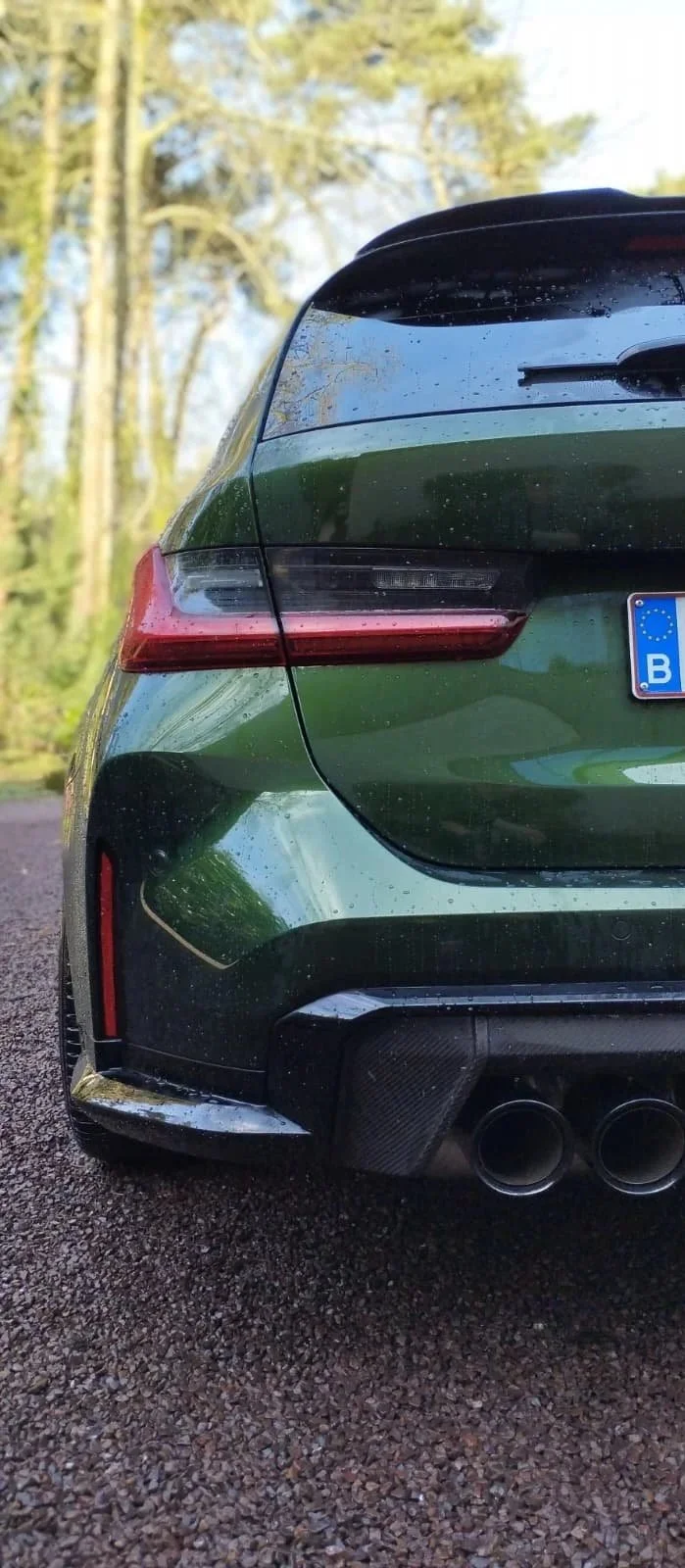 De achterkant van een groene auto met dubbele uitlaatpijpen, een aflineerbare spoiler en regen op de achterkant, geparkeerd op een paden in een bosgebied.