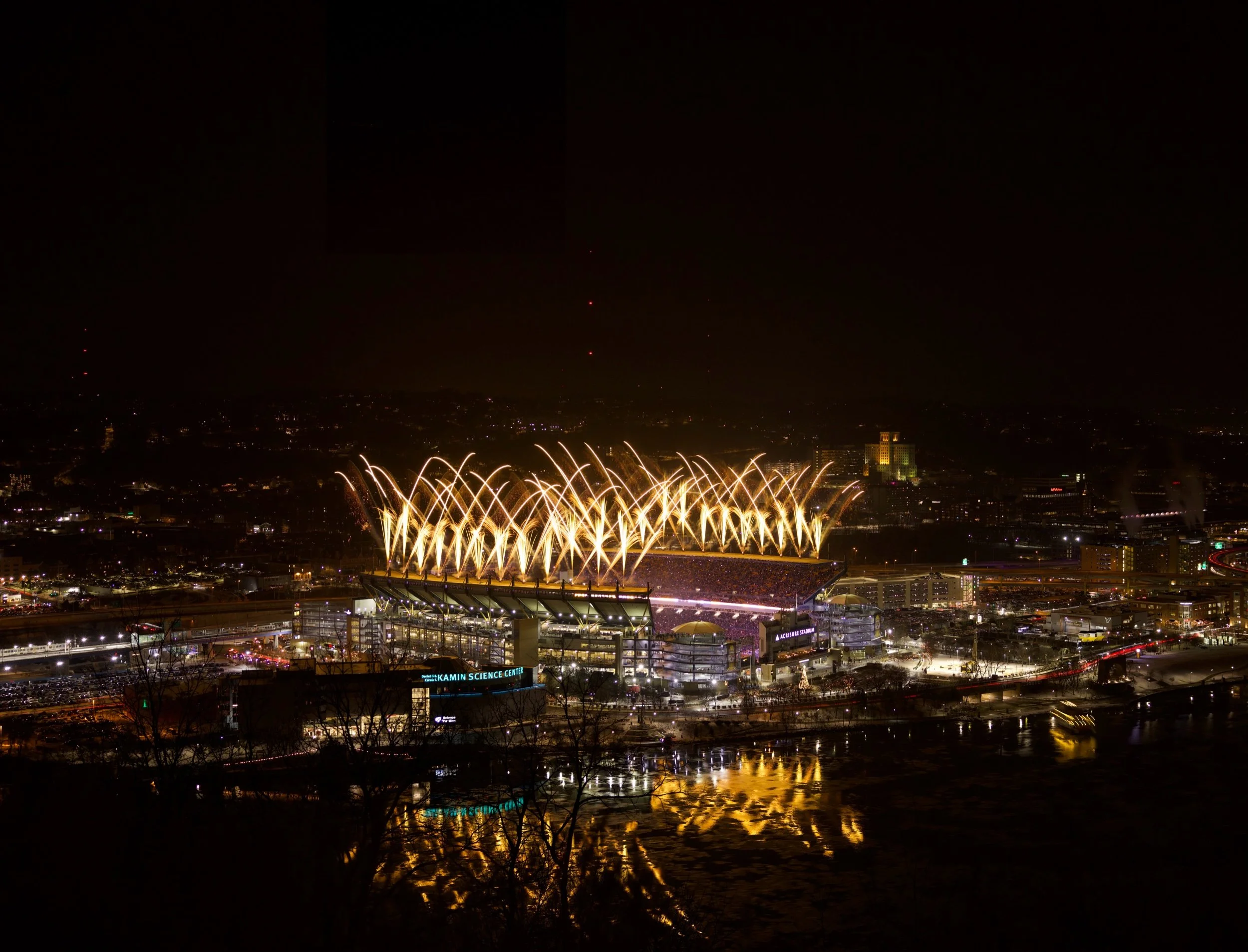 Nighttime view of a large stadium with fireworks illuminating the sky above, city lights reflecting on a nearby body of water.