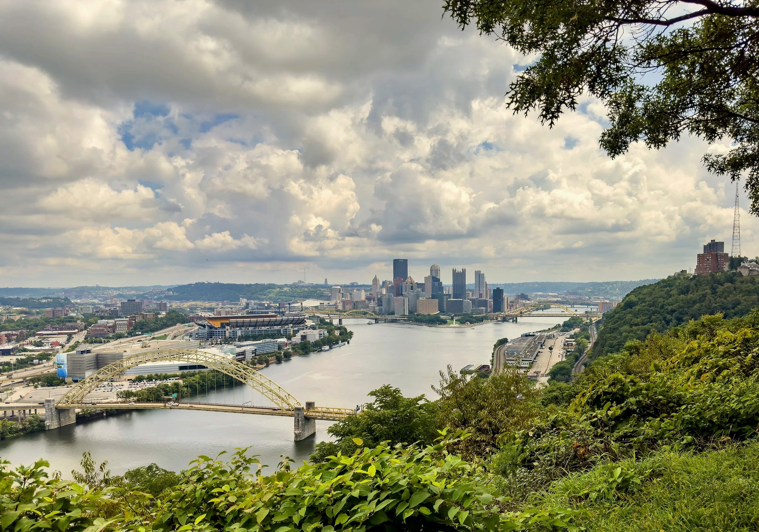 A panoramic view of Pittsburgh, Pennsylvania, with the city's skyline in the background, rivers, bridges, and green hills in the foreground under a partly cloudy sky.
