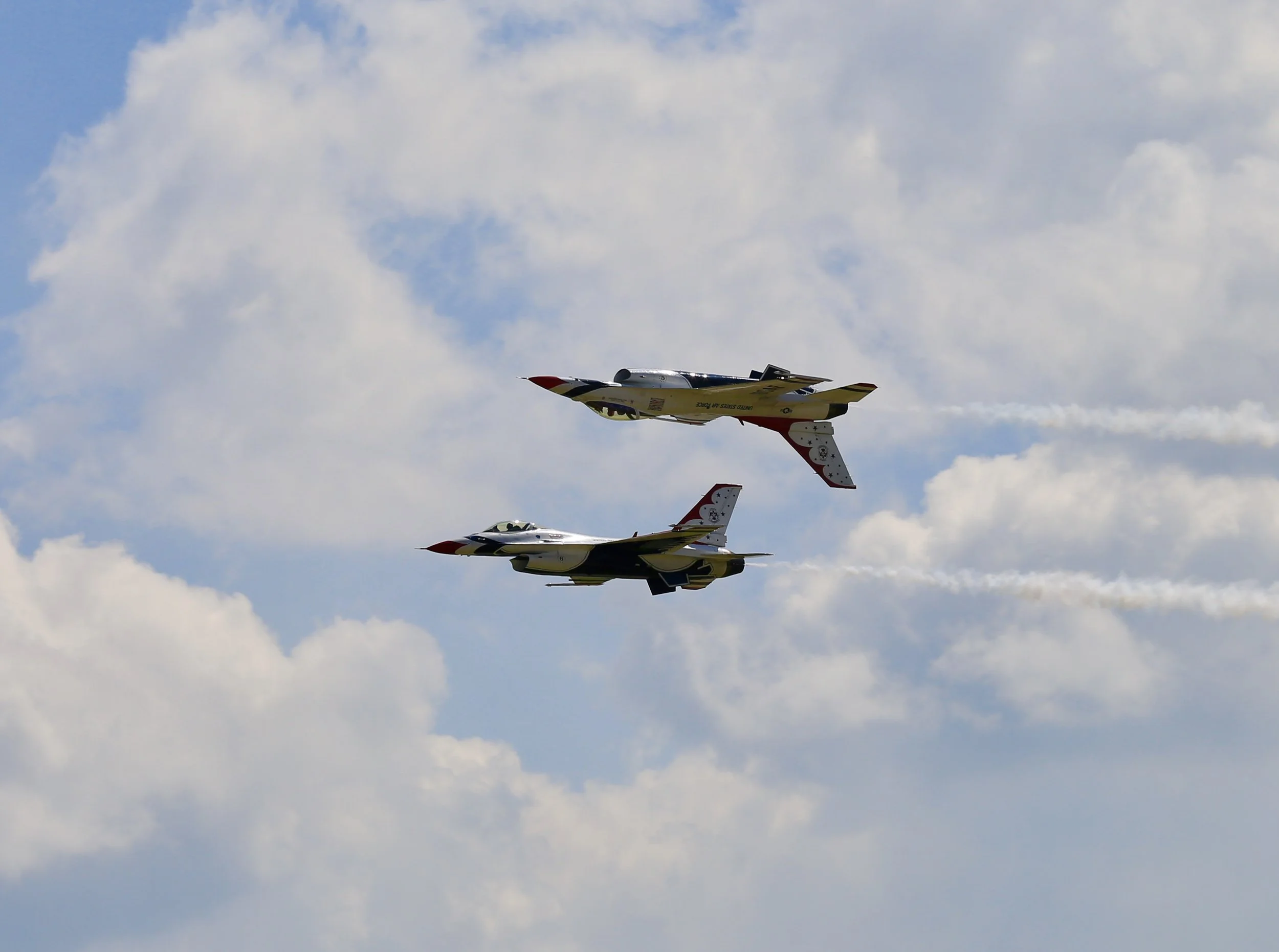 Two fighter jets flying in formation through a partly cloudy sky, leaving trails of vapor behind.