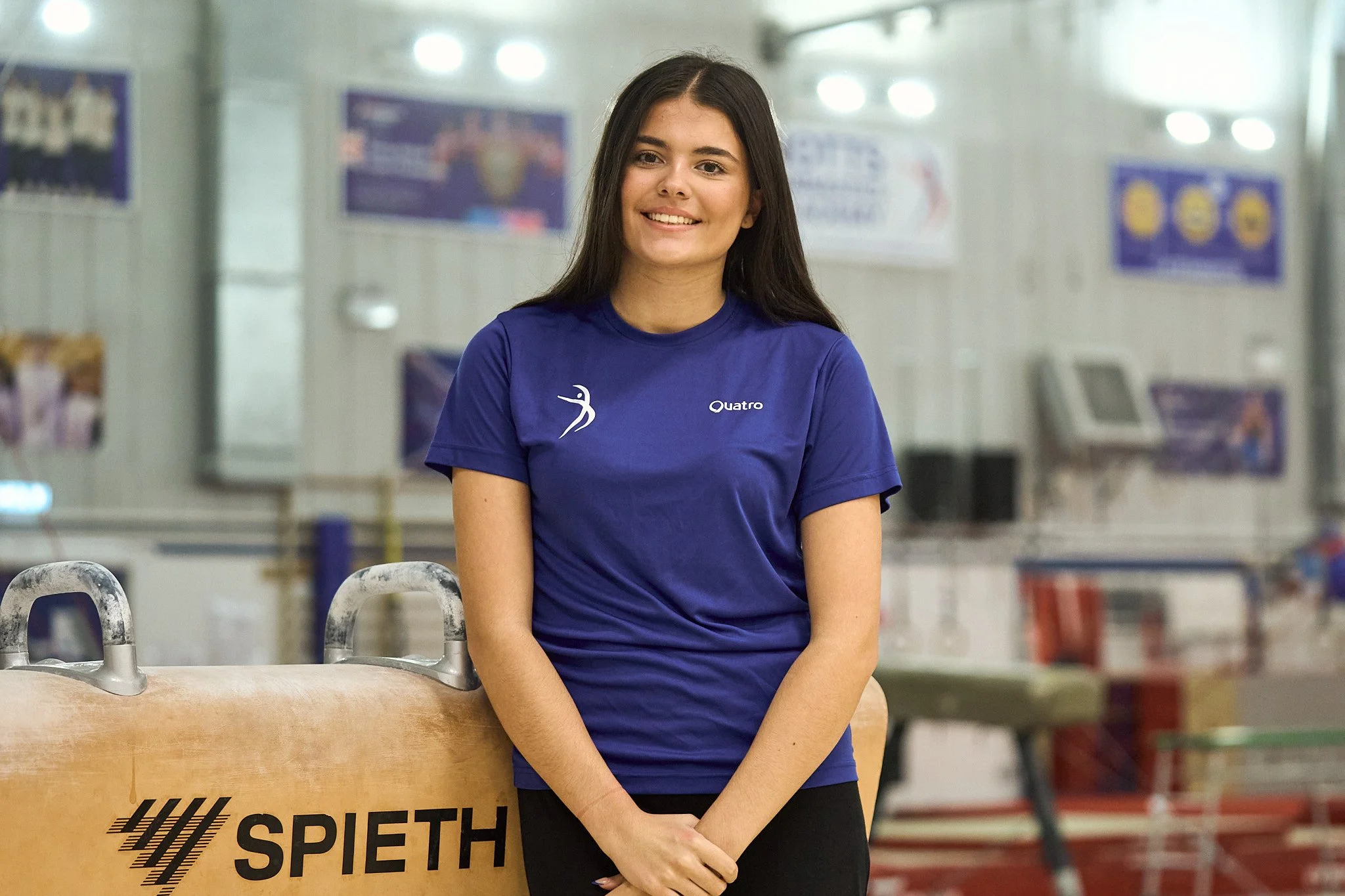 A young woman with long dark hair standing in an indoor sports facility, wearing a blue sports shirt and smiling at the camera. She is leaning against a wooden gymnastics vaulting table with the brand name "SPIETH" on it.