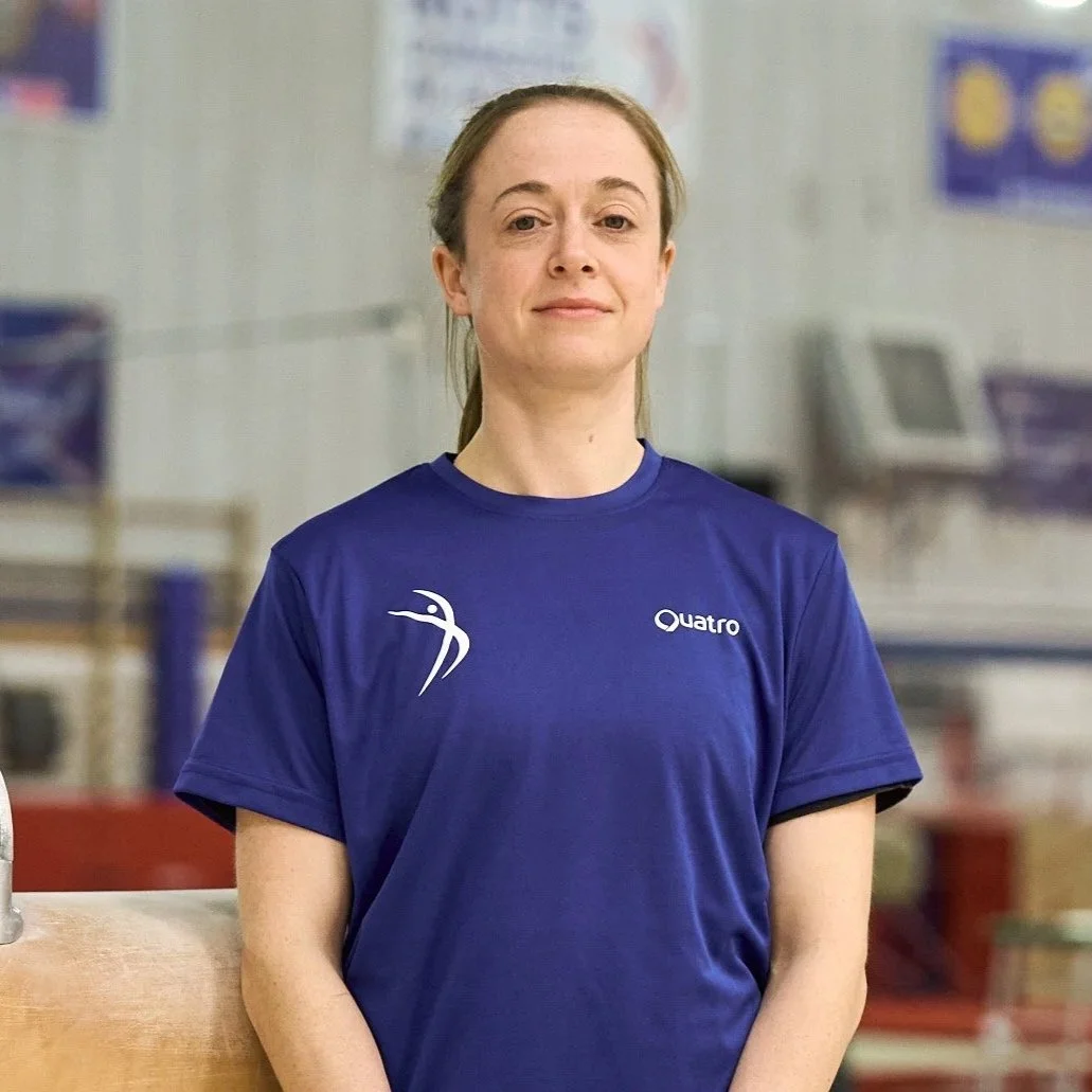 A woman in a blue athletic shirt standing indoors, smiling slightly, with a blurred background of gym equipment.