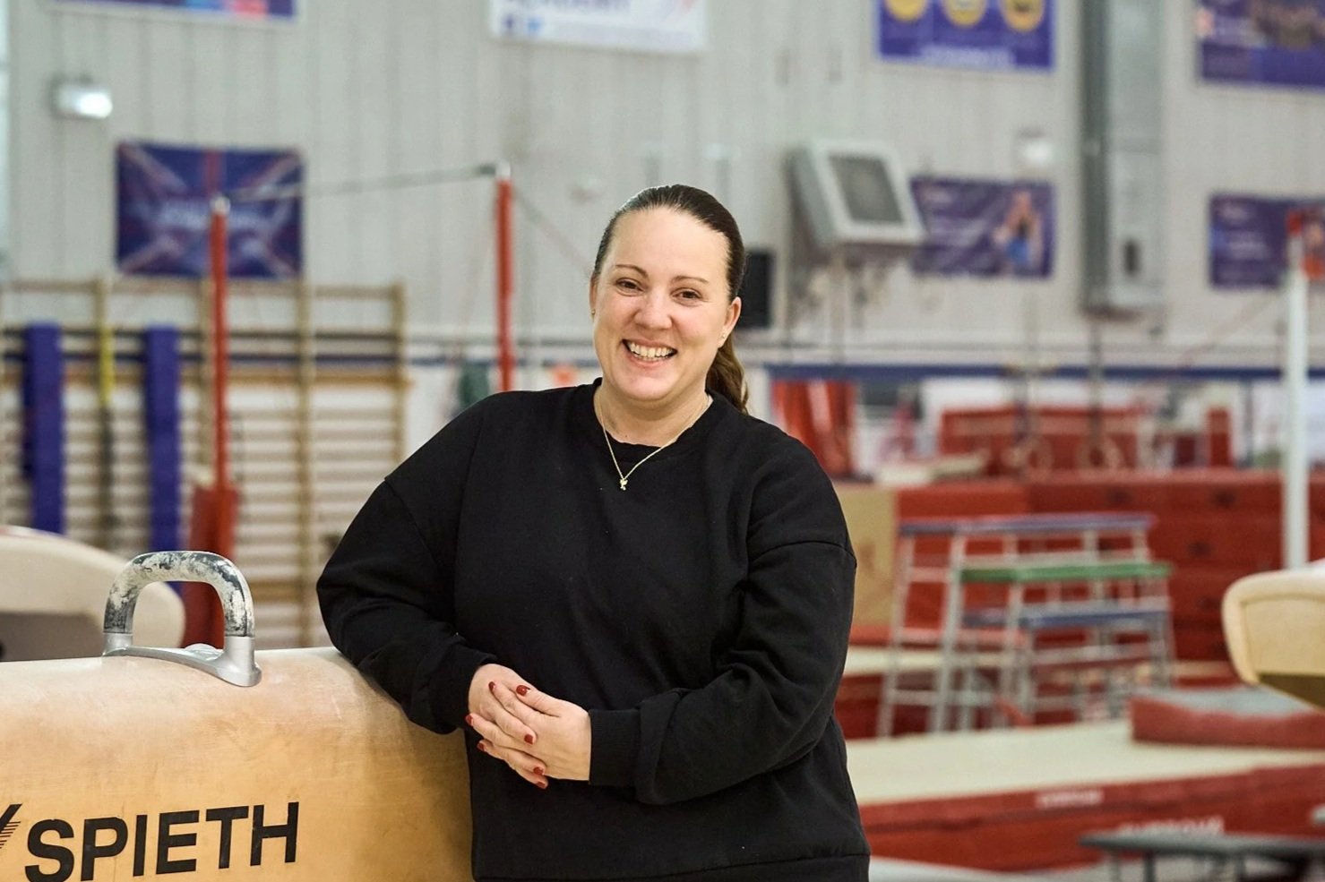 Close-up of a smiling woman with long light brown hair, wearing a dark top, standing against a plain white wall.