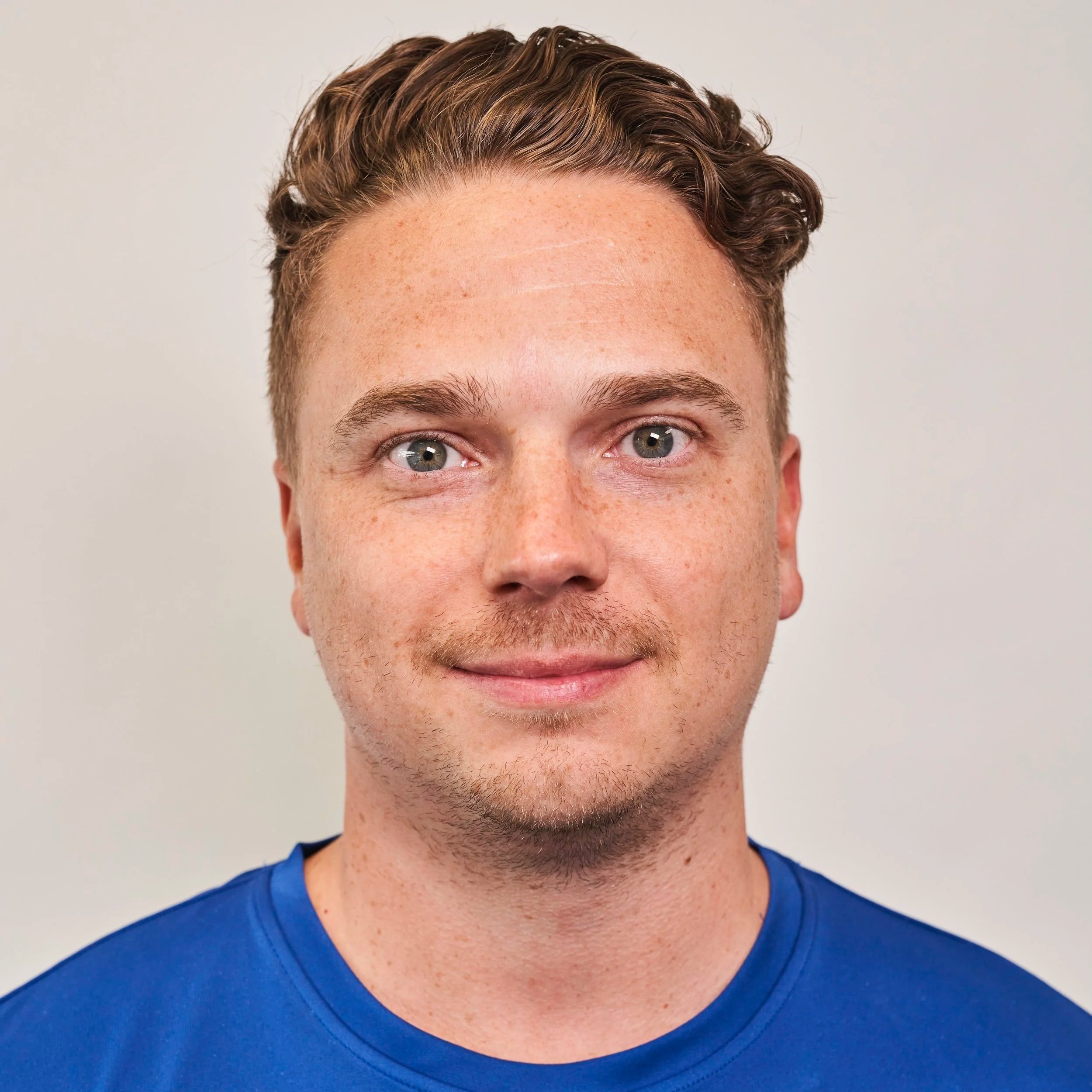 Close-up of a young man with light skin, short wavy brown hair, blue eyes, and freckles, wearing a blue shirt, smiling softly against a plain backdrop.