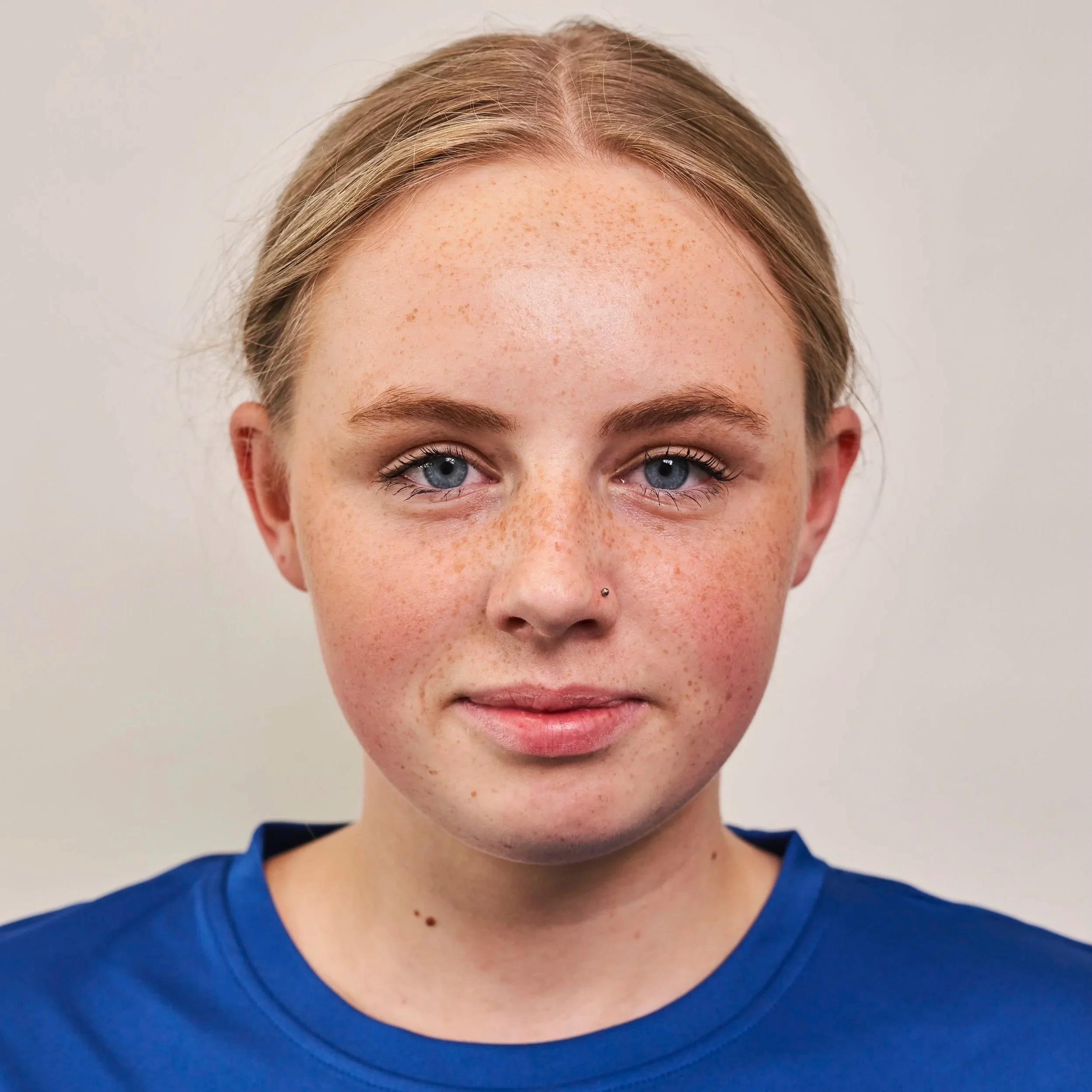 A close-up of a young woman with freckles, blue eyes, and light brown hair wearing a blue shirt, facing the camera with a neutral expression.