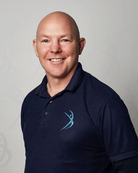 Portrait of a smiling man with a bald head wearing a navy blue polo shirt with a logo, standing against a plain gray background.