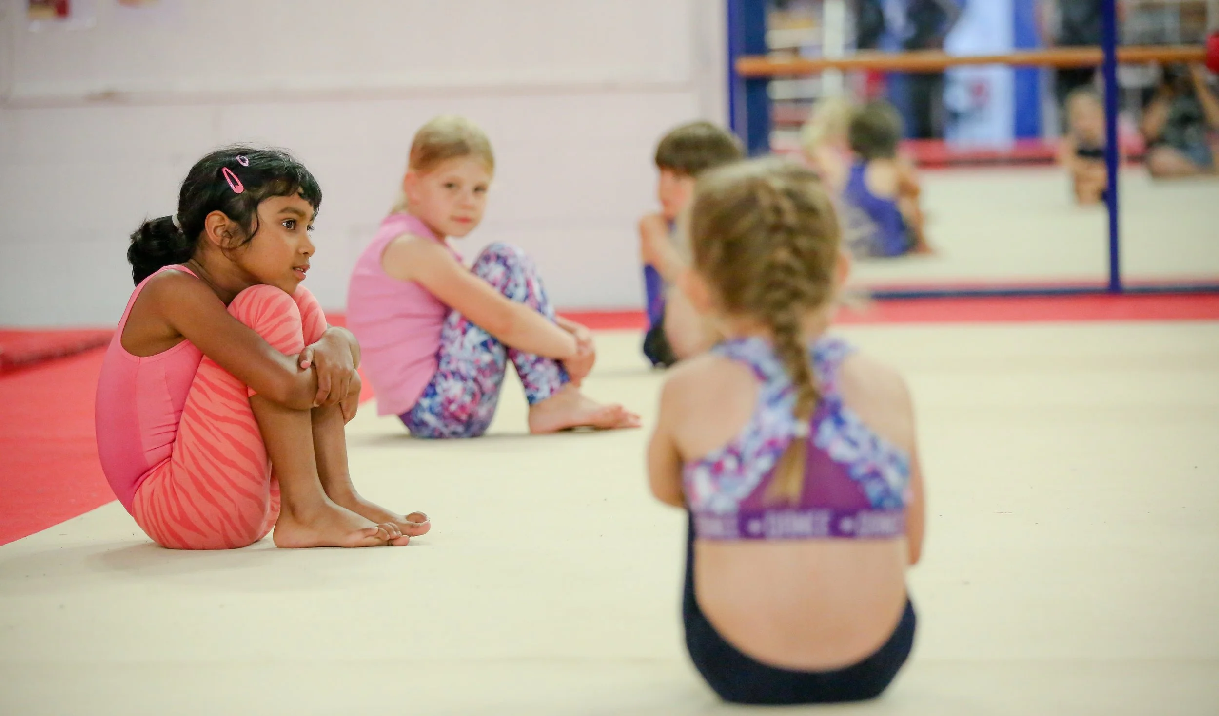 Children sitting on the floor of a gymnastics or dance studio, listening attentively, with a mirror and gym equipment in the background.