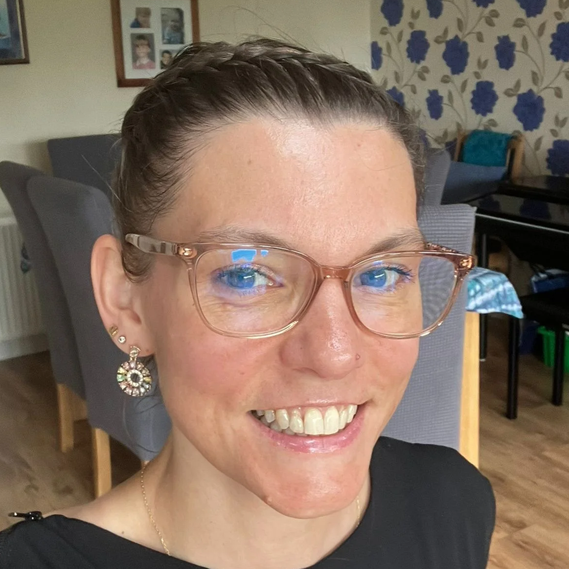Close-up of a woman with short wet hair, wearing glasses with a dog tag chain, earrings, and a black top, smiling in a home setting with dining table, chairs, and floral wallpaper in the background.