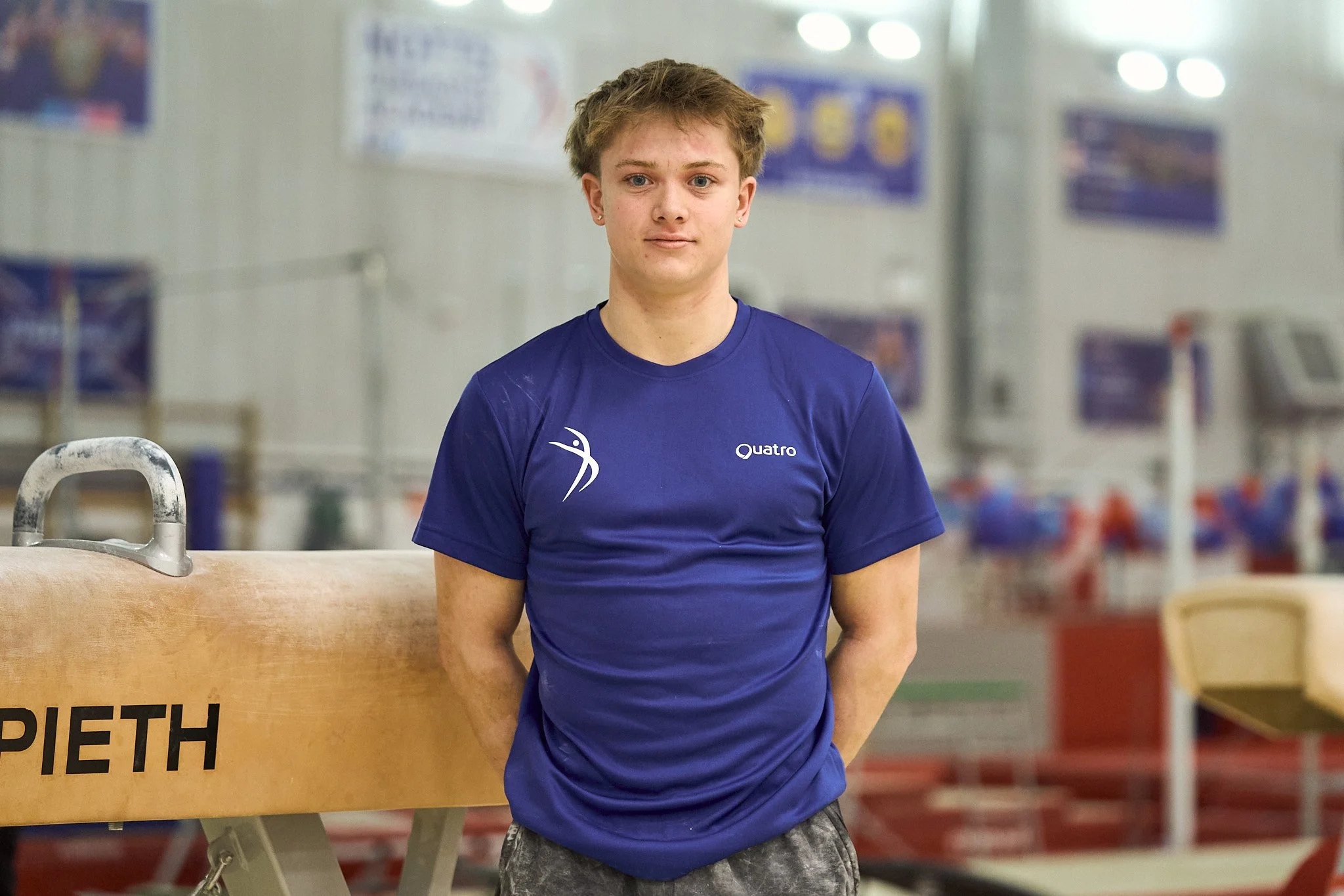 A young male athlete standing in a gym, wearing a blue athletic shirt with white logos, in front of gymnastics equipment, with a focused expression.