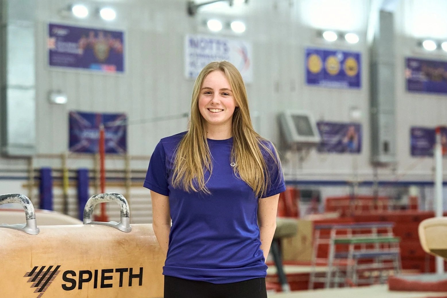 A close-up of a young woman with freckles, blue eyes, and light brown hair wearing a blue shirt, facing the camera with a neutral expression.