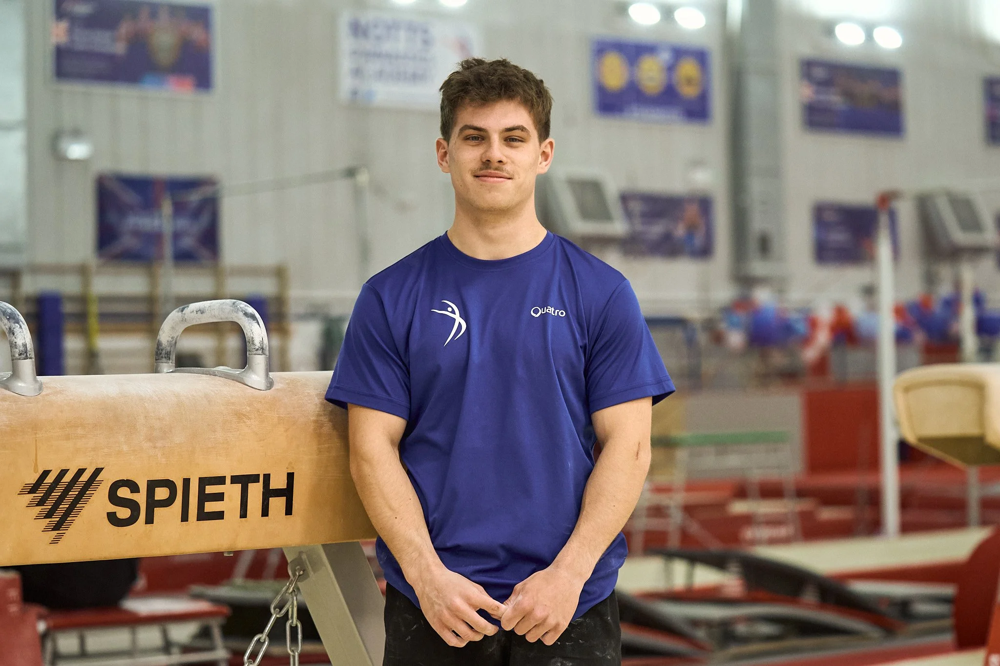A young male athlete with short brown hair and light skin stands in an indoor gym or sports facility, wearing a blue athletic shirt with logos, next to a wooden gymnastics pommel horse labeled 'SPIETH'.