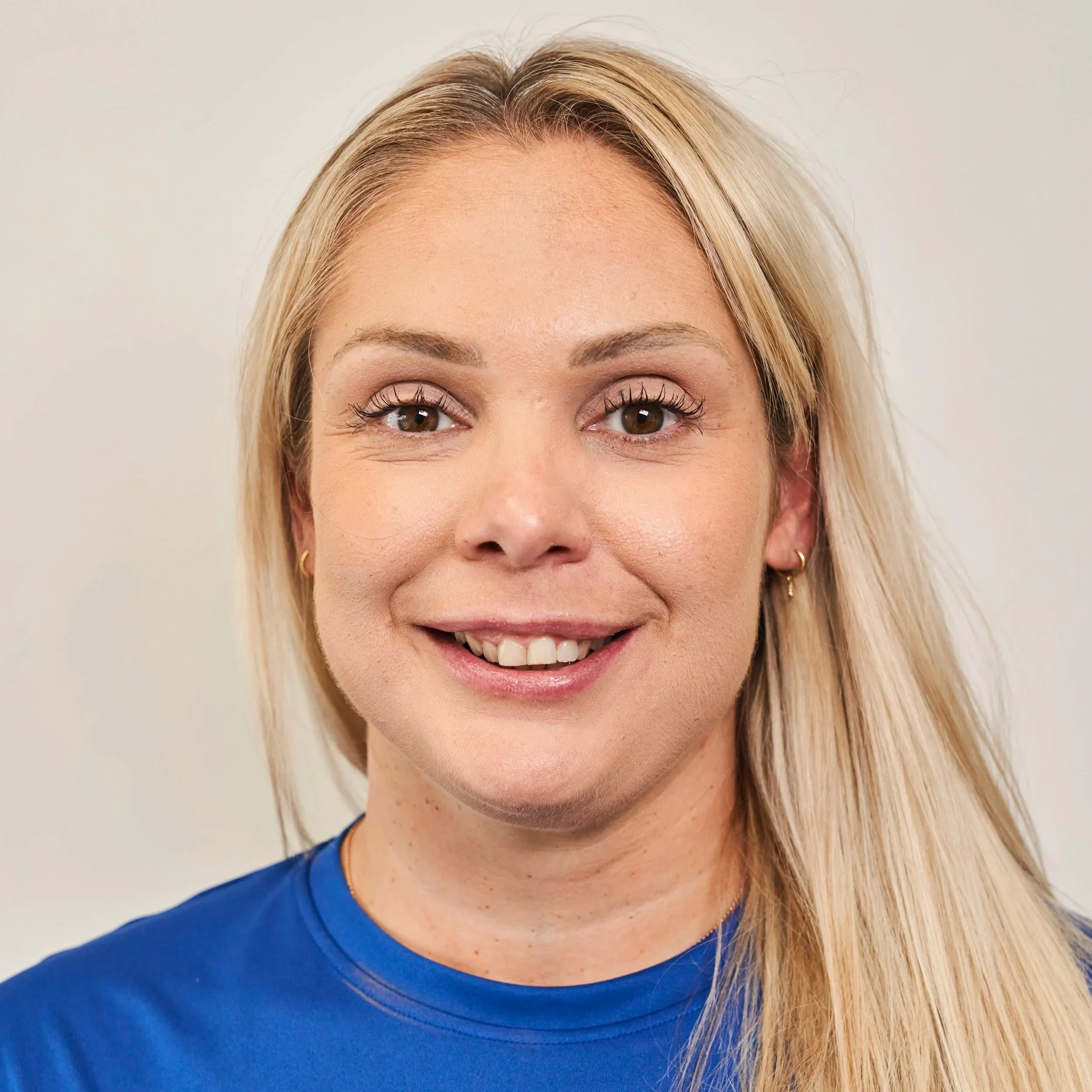 Close-up of smiling woman with long blonde hair, brown eyes, wearing a blue top and gold earrings.