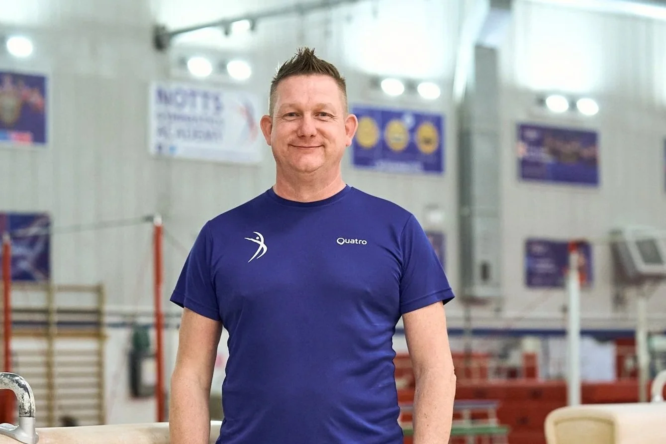 A man with short blond hair, wearing a white sports shirt with logos, smiling, against a plain light background.