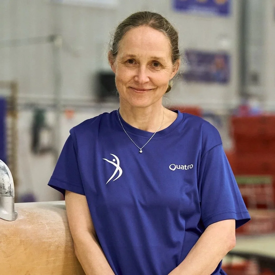 A smiling woman with gray hair wearing a blue sports shirt with a logo and the word 'Quatro' on it, standing in an indoor gym or sports facility.
