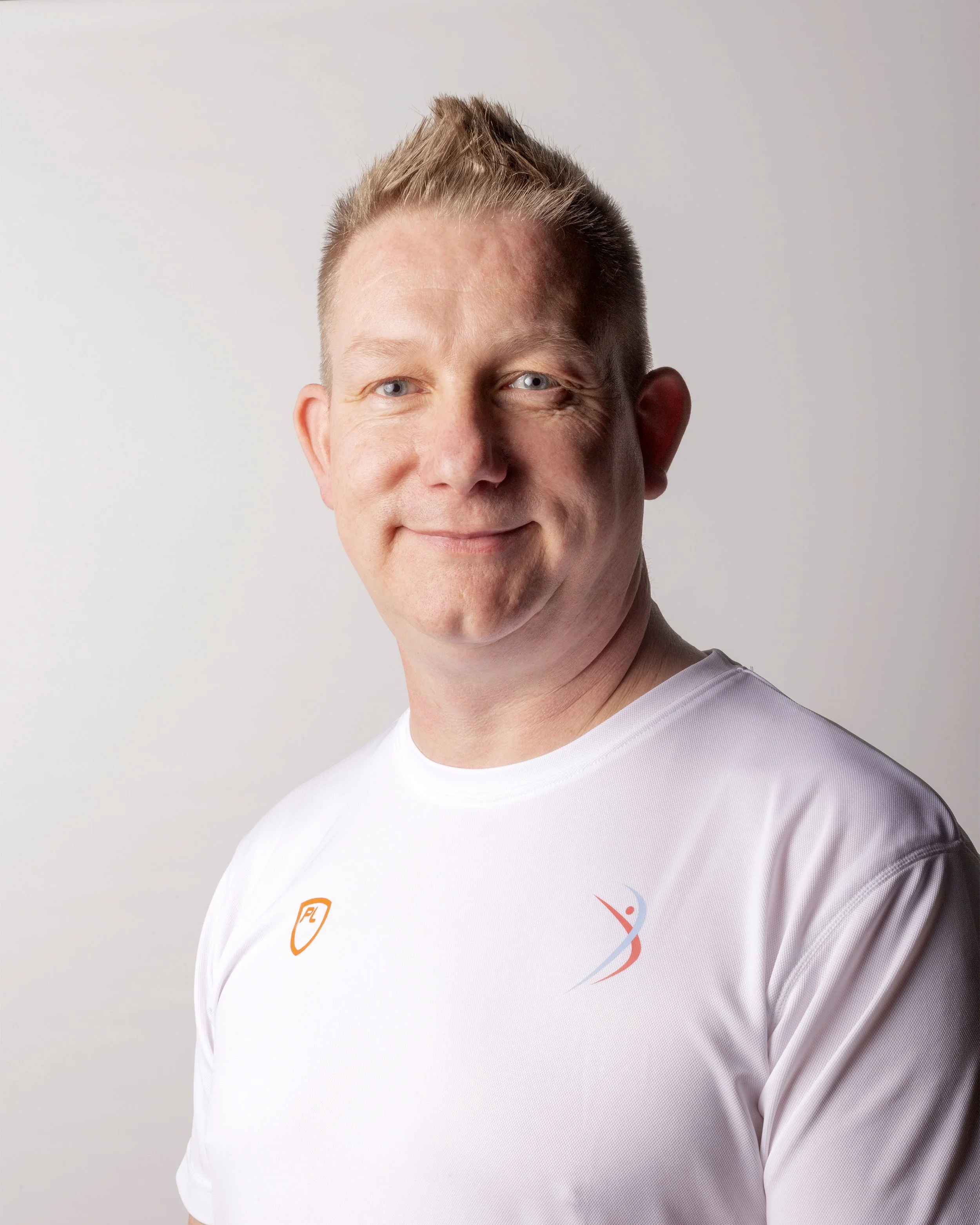 Portrait of a smiling man with short blonde hair wearing a white athletic shirt with logos on the chest against a plain light background.