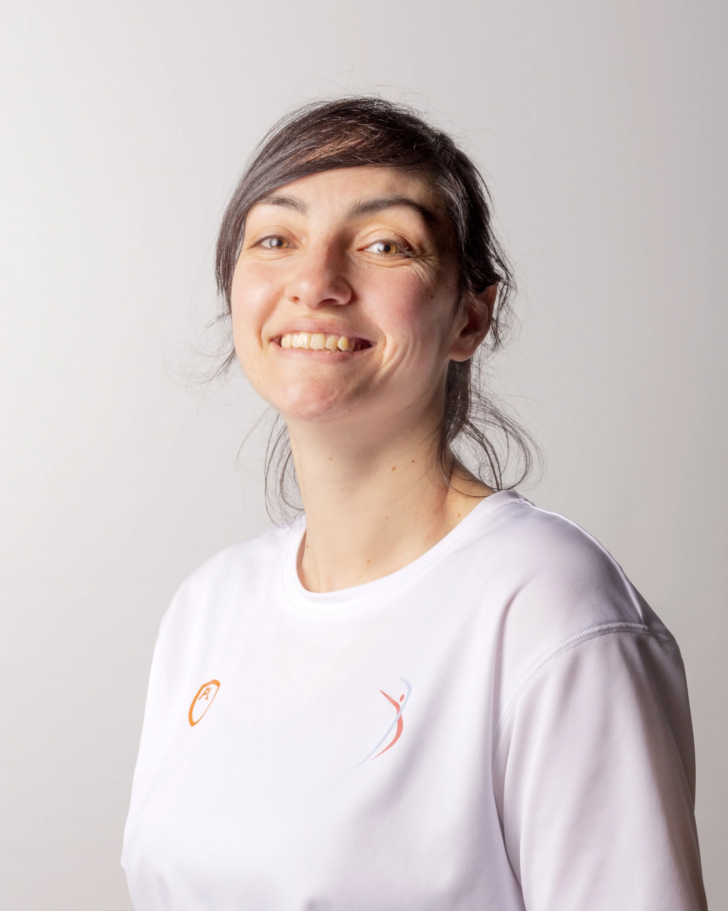 A smiling woman with short brown hair and light skin, wearing a white t-shirt with a logo on the chest, standing against a plain light gray background.