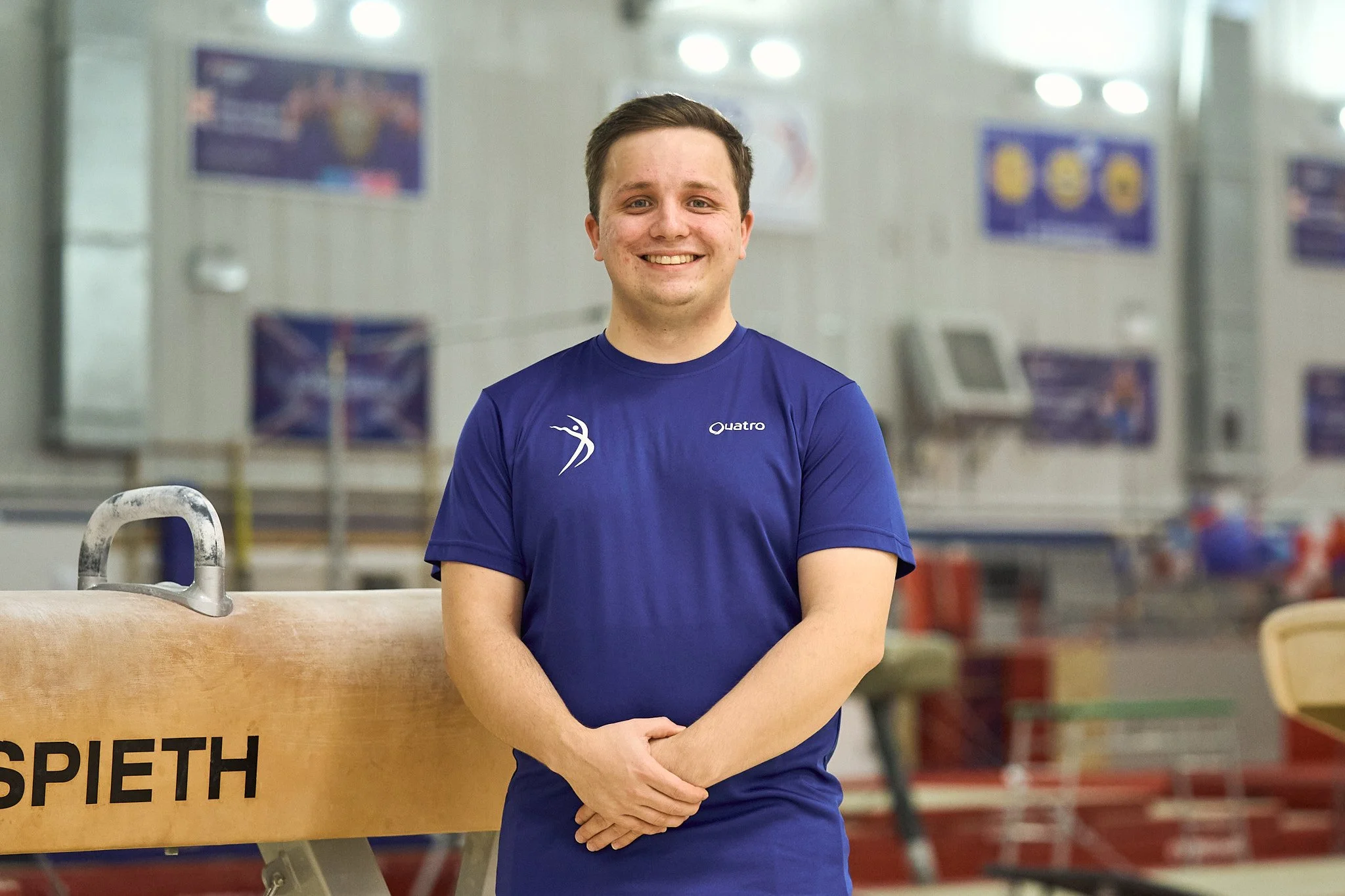 A young man with short brown hair wearing a blue athletic t-shirt stands in a gymnasium, smiling at the camera. There is a wooden gymnastics pommel horse with