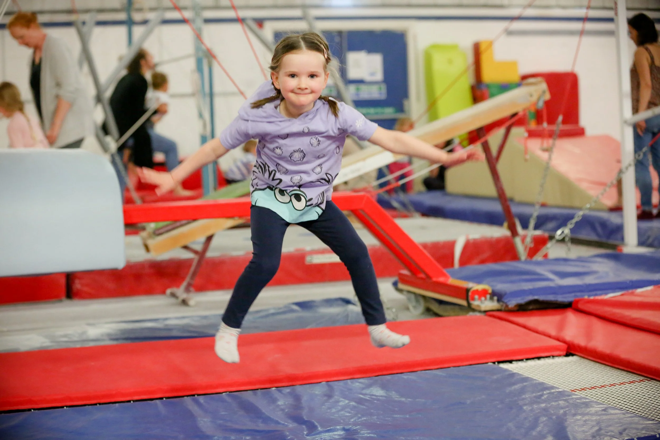 Young girl in a purple shirt and dark pants jumping on a trampoline at an indoor trampoline park with other children and adults in the background.