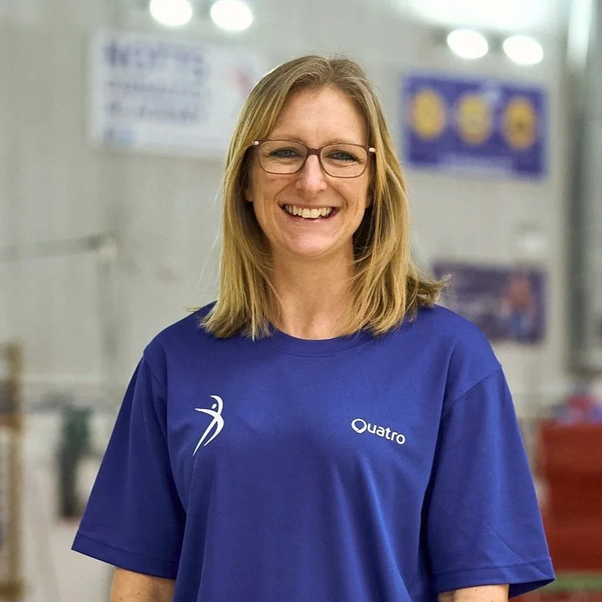 A smiling woman with shoulder-length blonde hair and glasses, wearing a blue Quatro sports shirt, standing indoors with blurred posters in the background.
