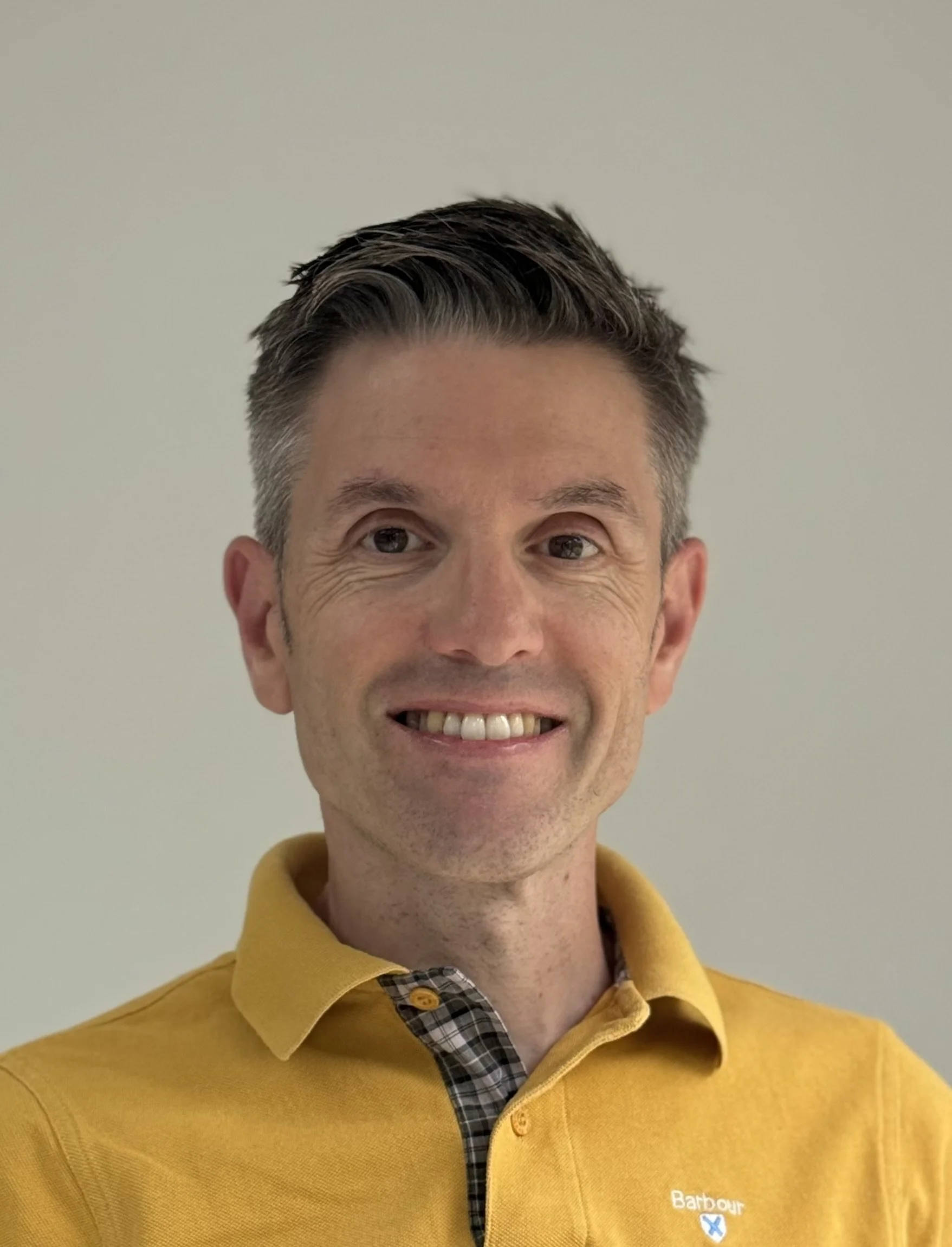 A smiling man with short dark hair, wearing a yellow polo shirt with a plaid collar, stands against a plain beige background.