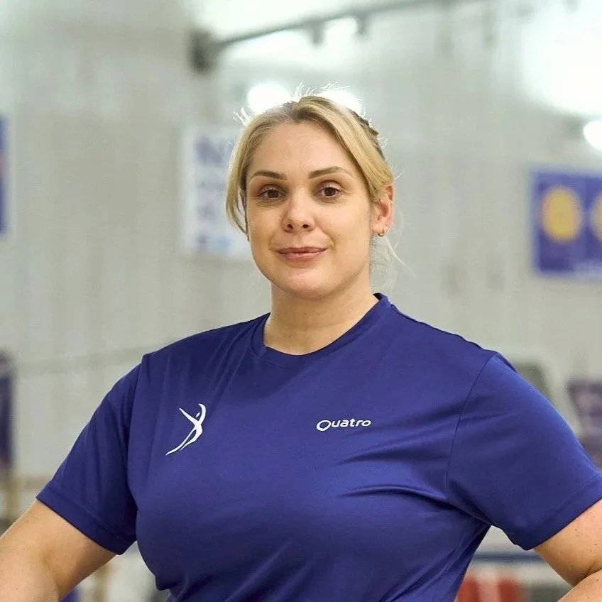 A woman with blonde hair tied back, wearing a blue athletic shirt with the words 'Quatro' and a logo, standing in a gym.