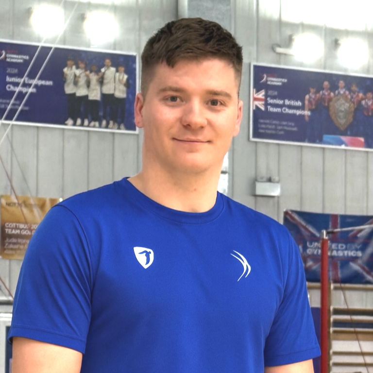 Portrait of a young man with short, tousled reddish-brown hair, blue eyes, and fair skin, wearing a blue shirt, smiling softly at the camera.