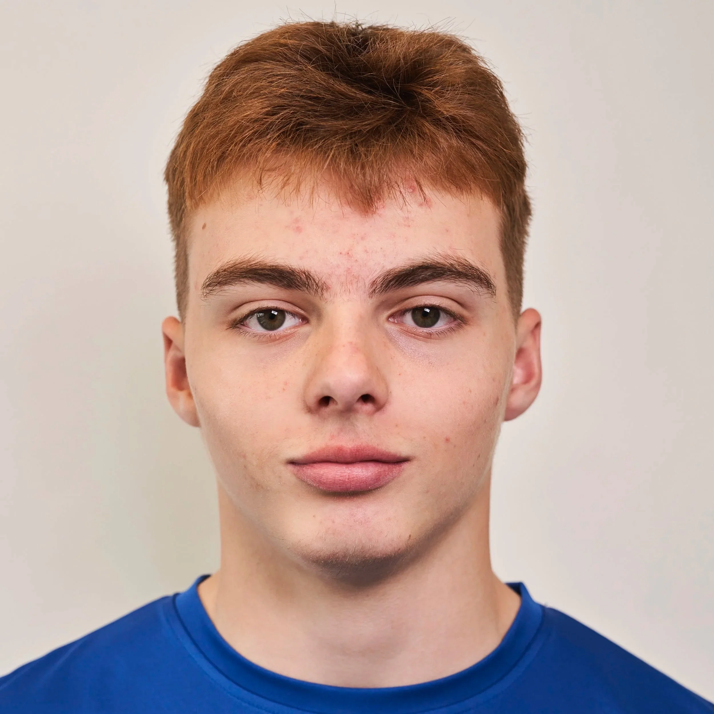 Portrait of a young man with red hair, wearing a blue shirt, facing the camera against a plain background.