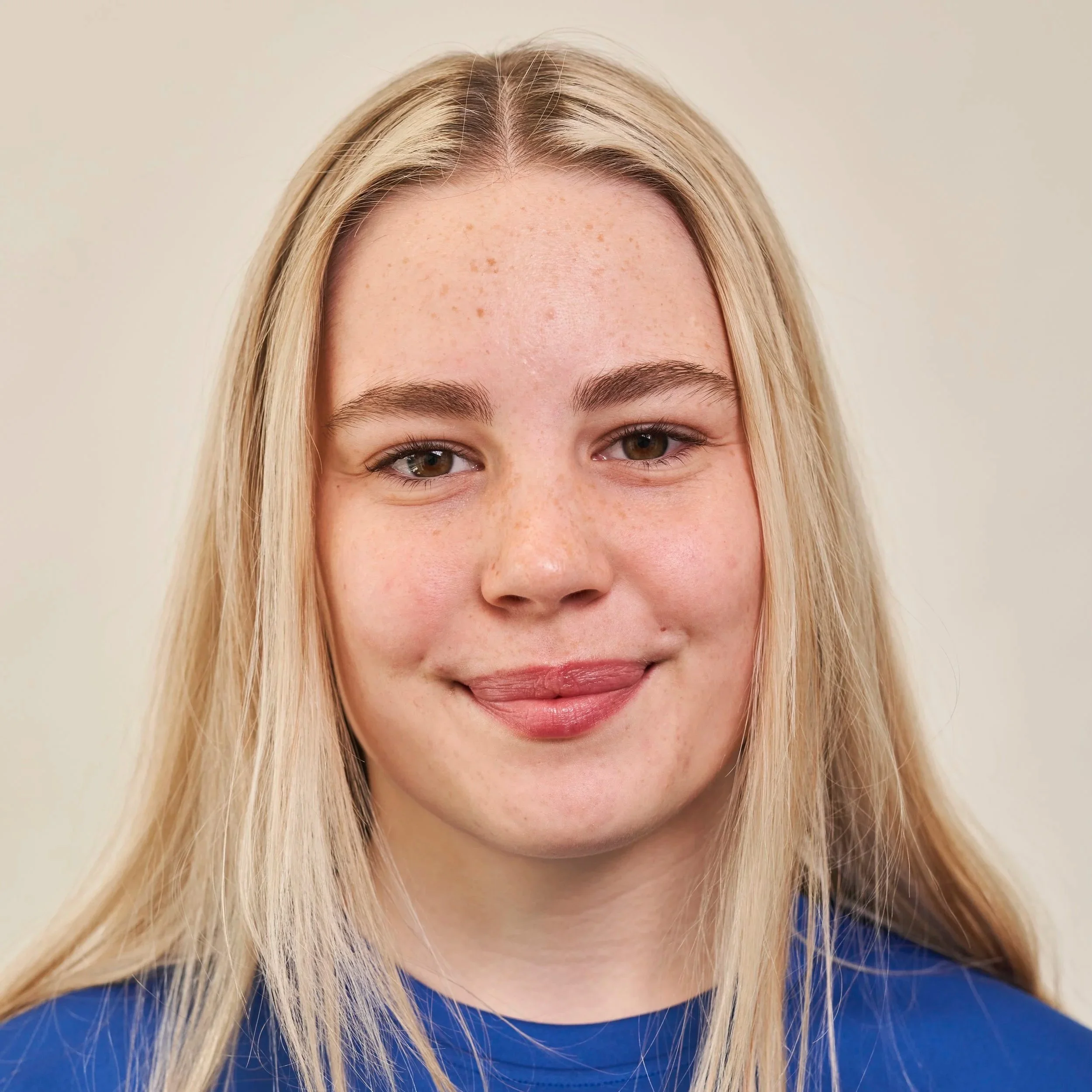 Close-up of a young woman with long blonde hair, light skin, and freckles, smiling in front of a plain background.