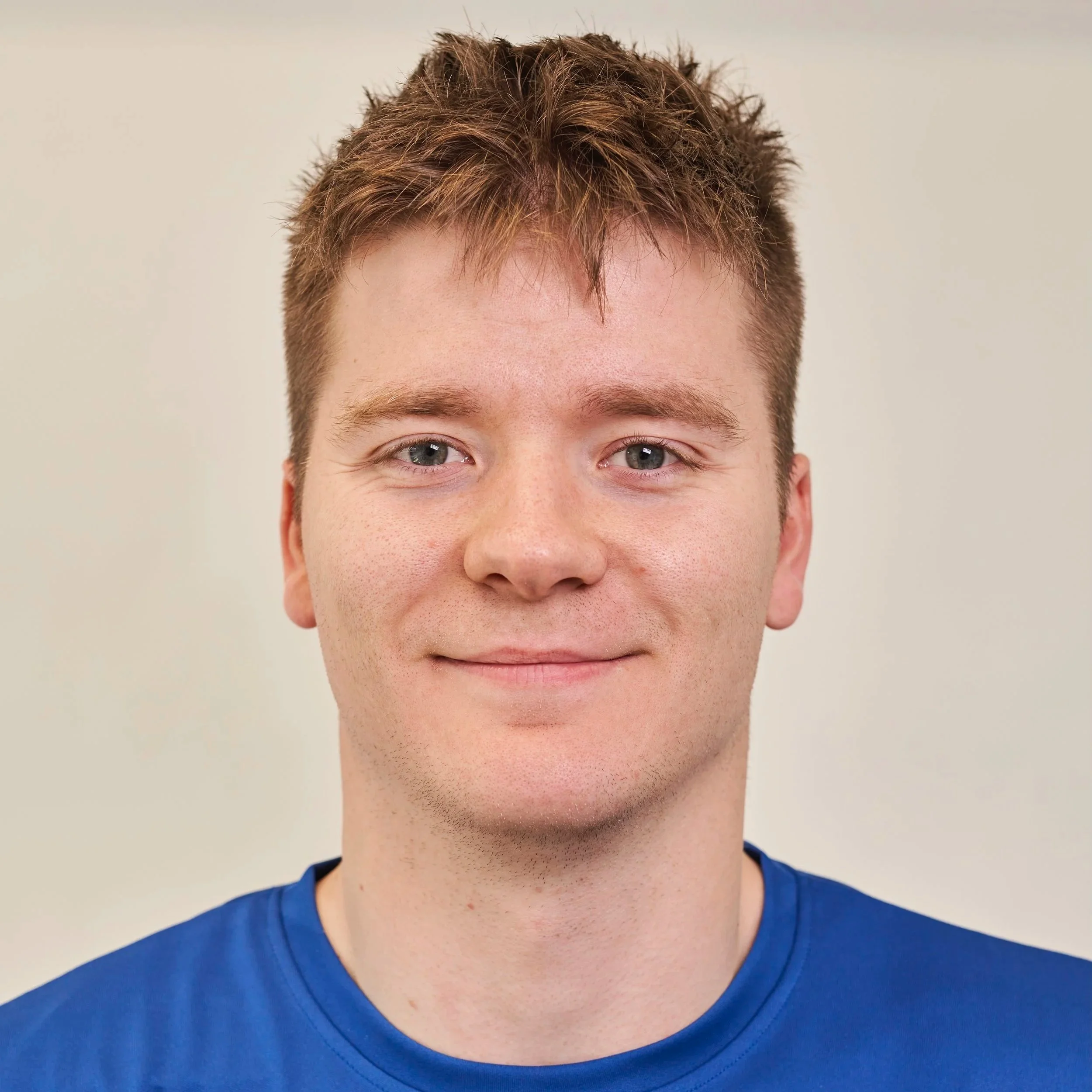 Portrait of a young man with short, tousled reddish-brown hair, blue eyes, and fair skin, wearing a blue shirt, smiling softly at the camera.