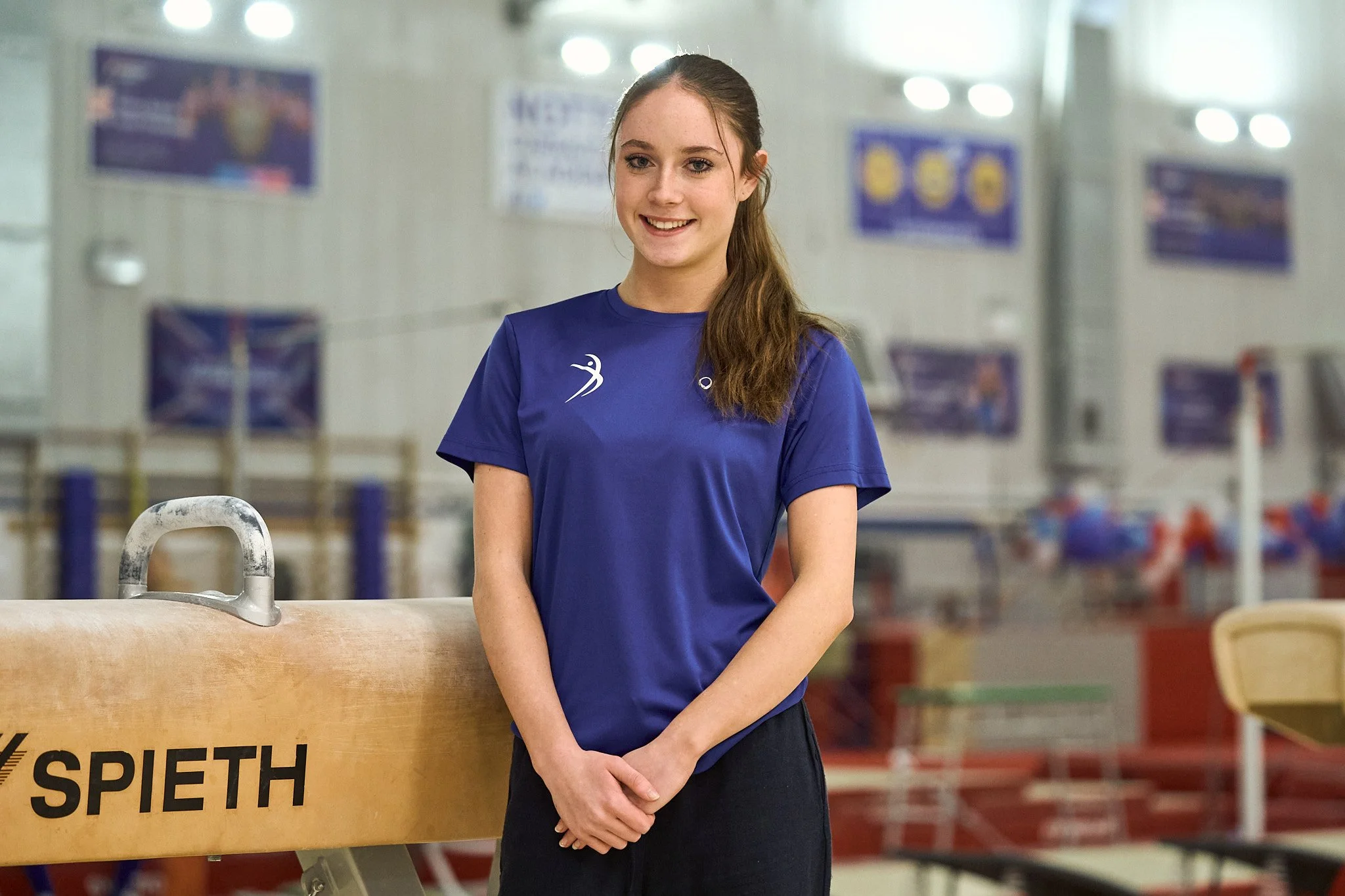 Young woman with long brown hair in a ponytail, wearing a blue sports shirt with white logos, standing in an indoor gymnastics training facility. She is smiling and standing next to a gymnastics springboard labeled SPIETH.