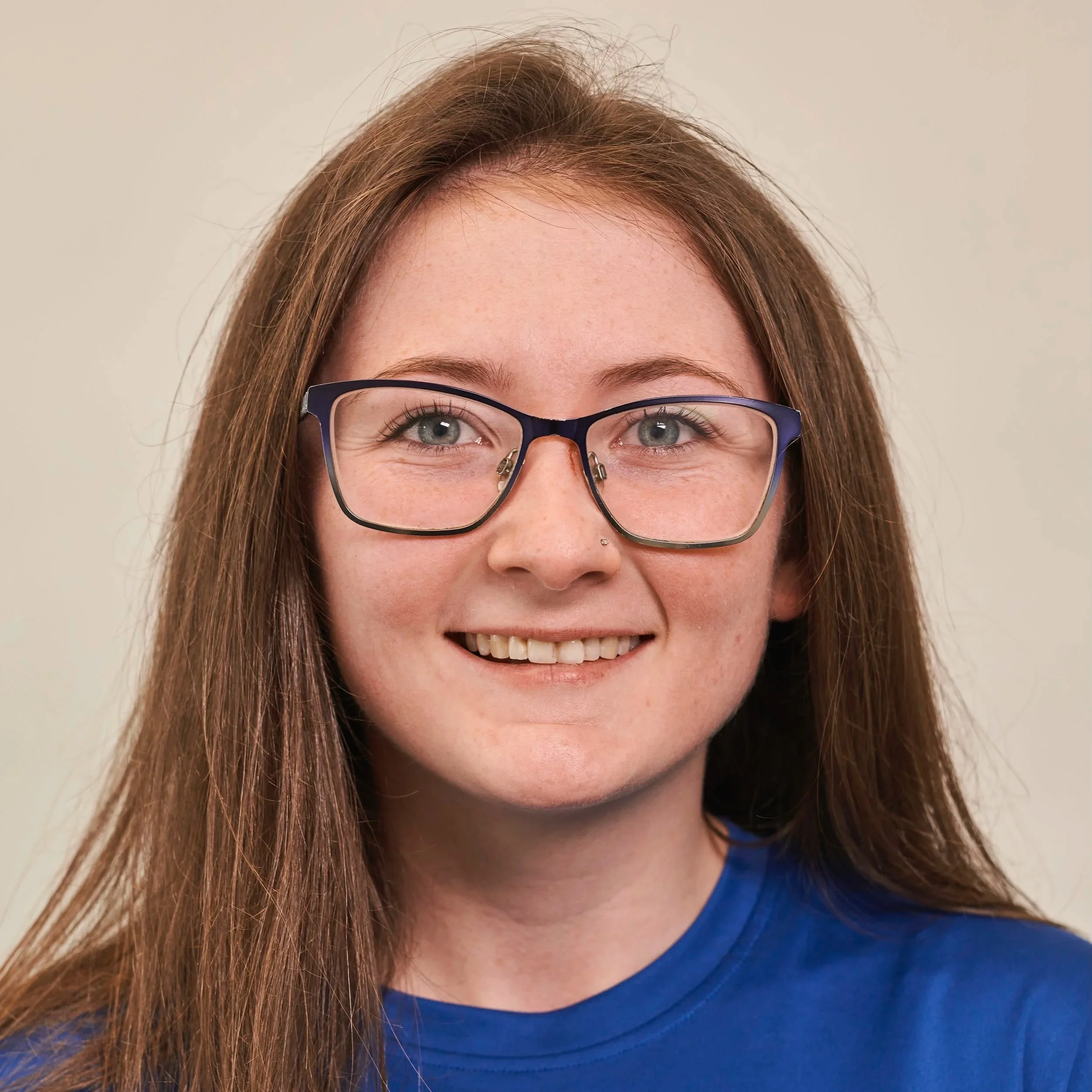A young woman with long brown hair, glasses, and a nose piercing, smiling at the camera, wearing a blue shirt.