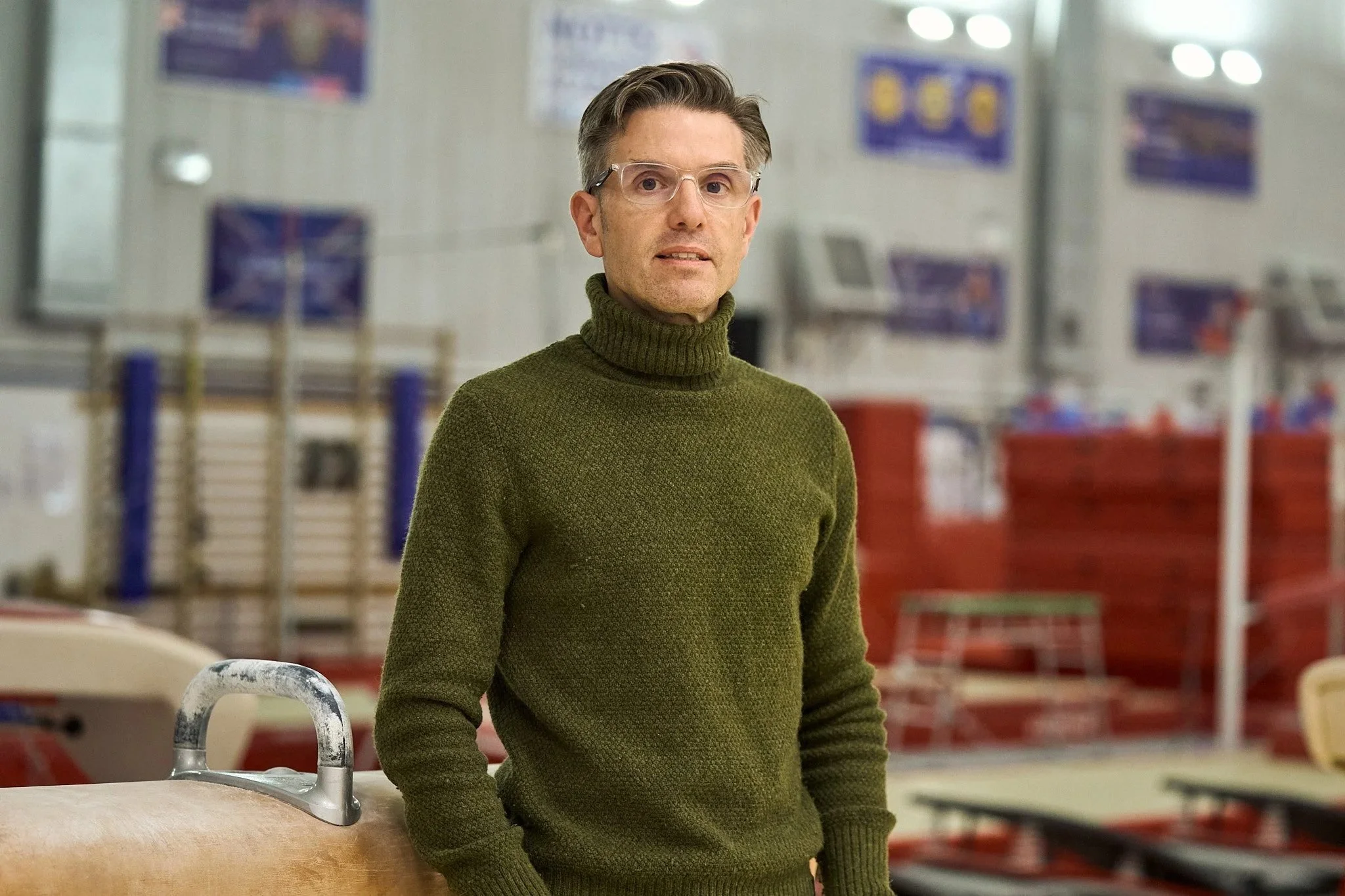 A smiling man with short dark hair, wearing a yellow polo shirt with a plaid collar, stands against a plain beige background.