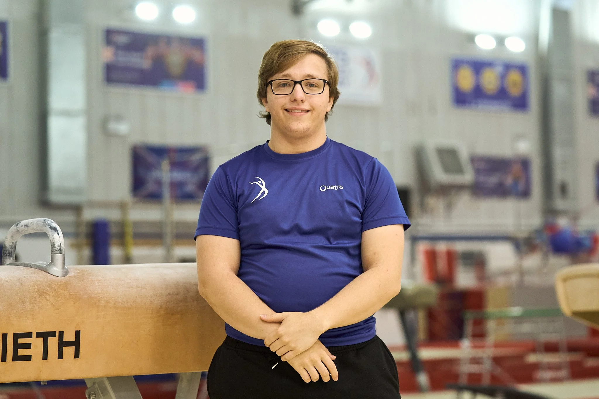 Young man with glasses and brown hair, smiling, wearing a blue athletic shirt, standing in an indoor sports facility with gymnastics equipment.