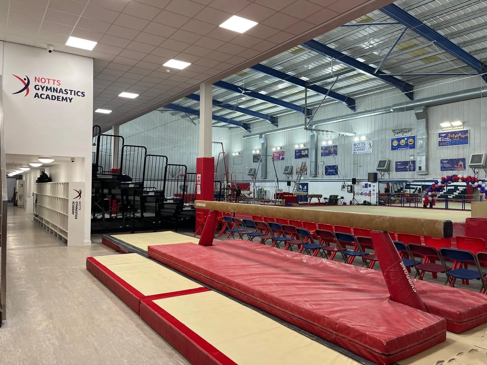 Interior of a gymnastics training center featuring a springboard, mats, and a vaulting apparatus. There are chairs arranged for spectators and banners on the walls.