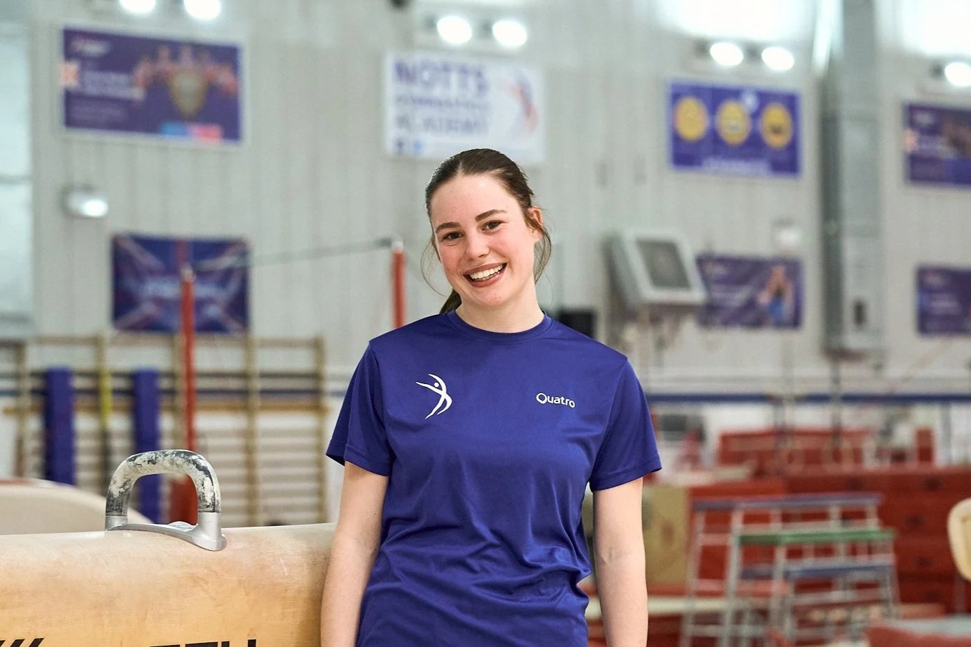 Close-up of a young woman with long blonde hair, light skin, and freckles, smiling in front of a plain background.
