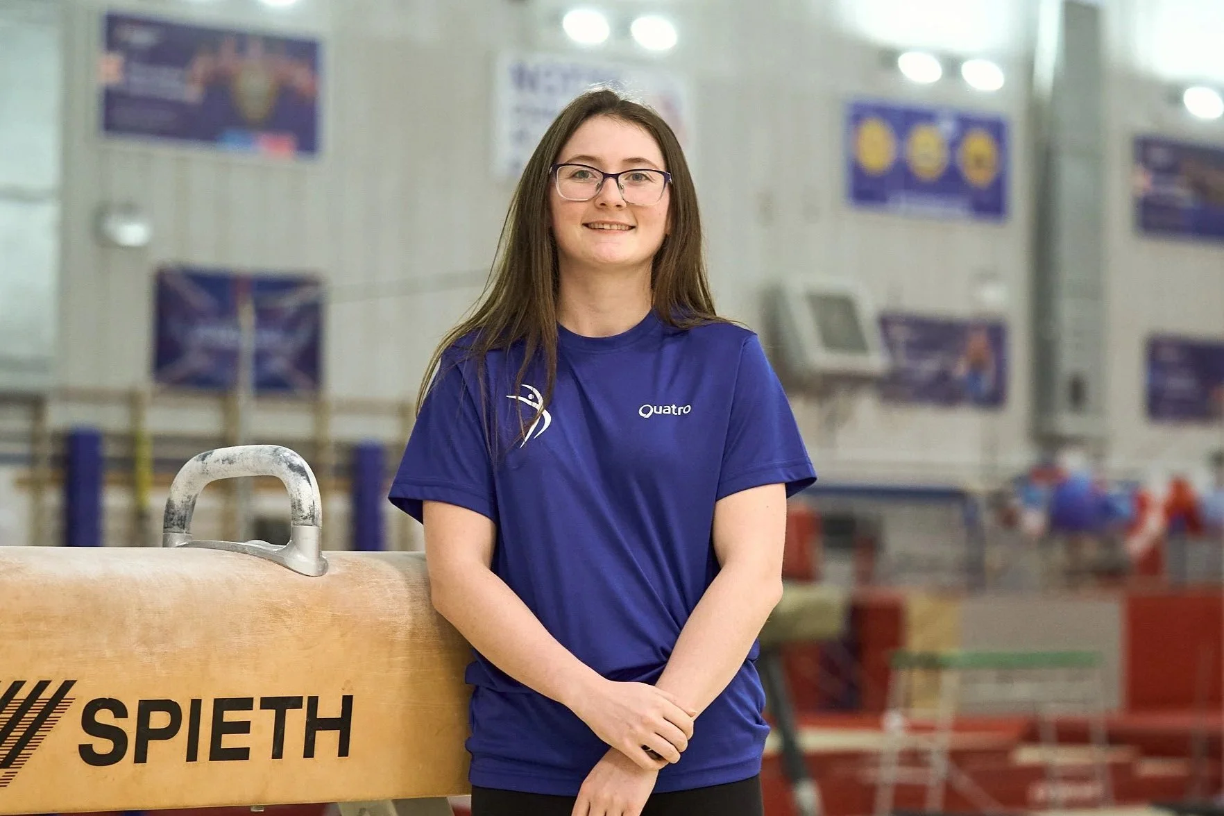 A young woman with long brown hair, glasses, and a nose piercing, smiling at the camera, wearing a blue shirt.
