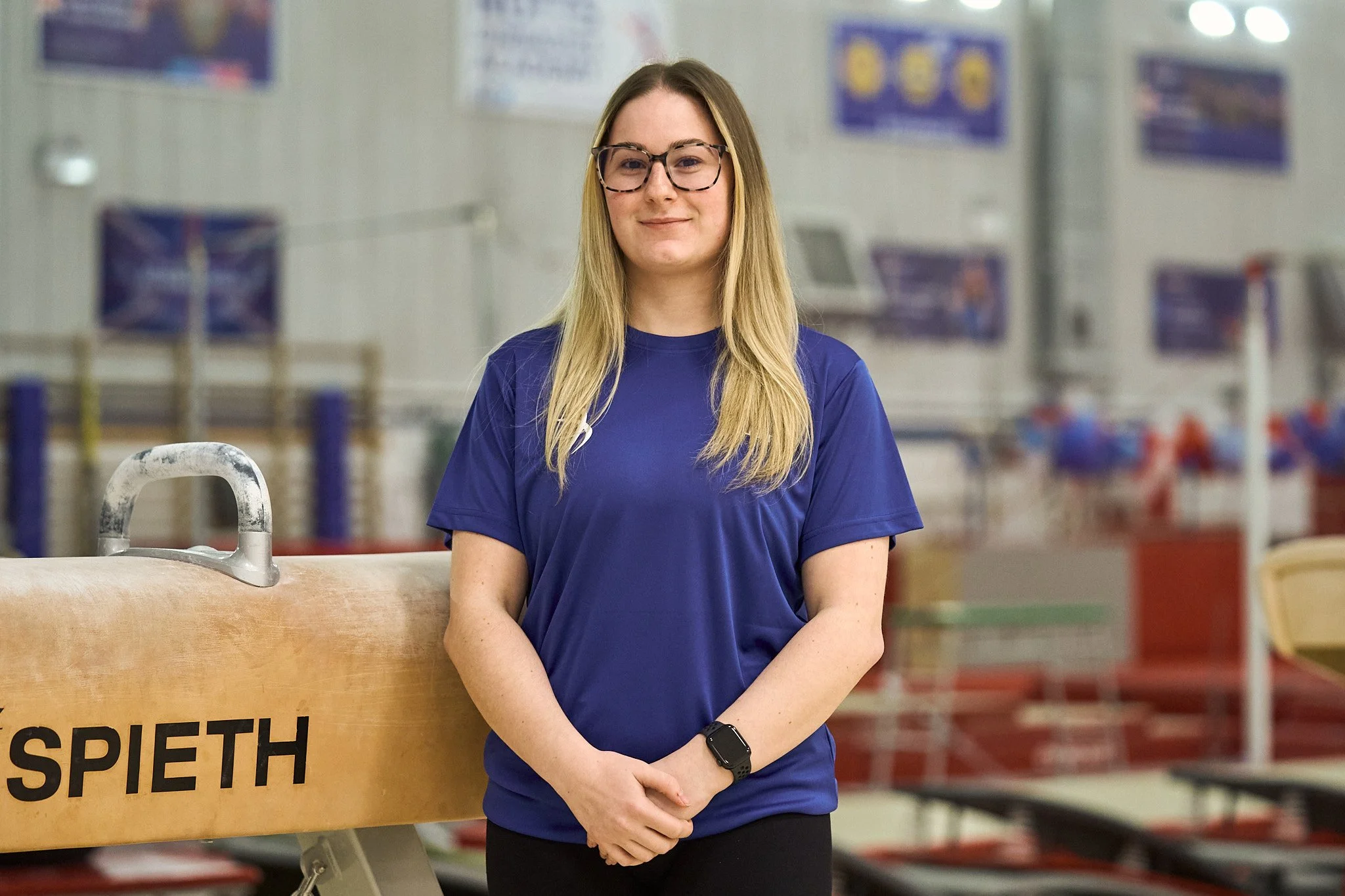 A young woman with long blonde hair and glasses standing in an indoor sports facility, wearing a blue athletic shirt and a black smartwatch, smiling softly with her hands clasped in front of her.
