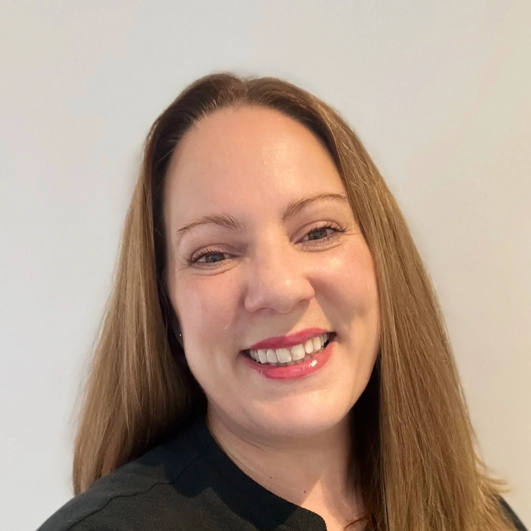Close-up of a woman with long, light brown hair, smiling, wearing a dark top, against a plain light-colored background.