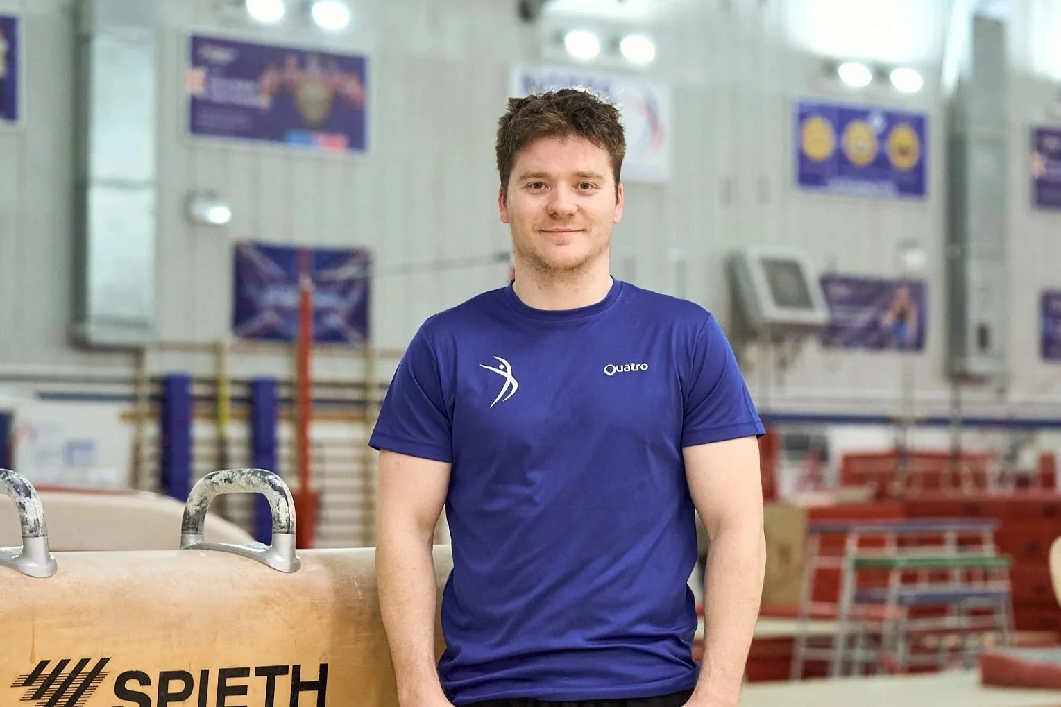 Portrait of a young man with short, tousled reddish-brown hair, blue eyes, and fair skin, wearing a blue shirt, smiling softly at the camera.