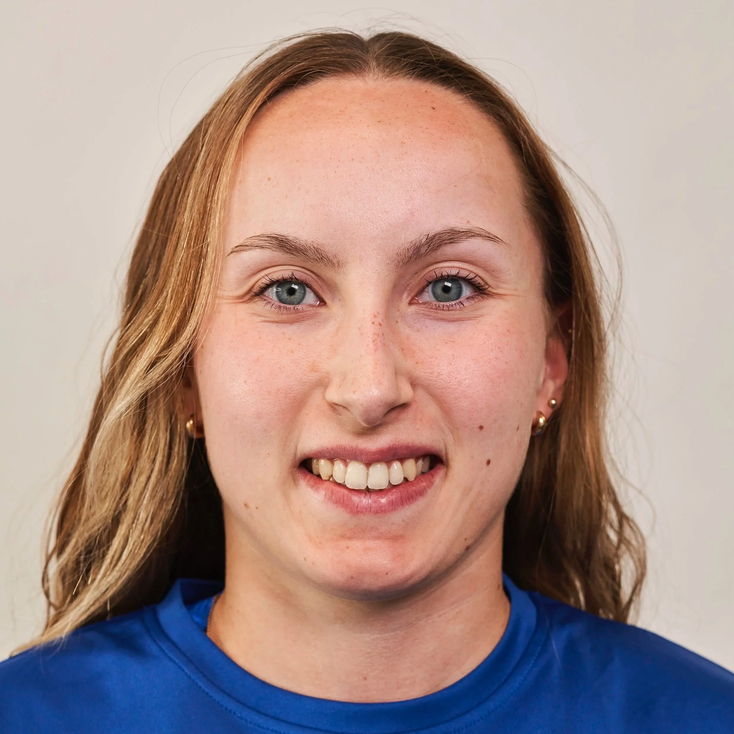 Close-up of a young woman with blue eyes and wavy brown hair, smiling, wearing small gold earrings and a blue sports shirt.