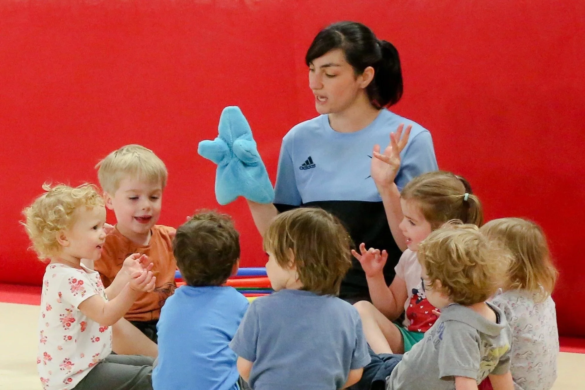 A woman dressed as a blue shark puppet interacts with a group of young children sitting on the floor, engaging them in a story or activity, with a red background.