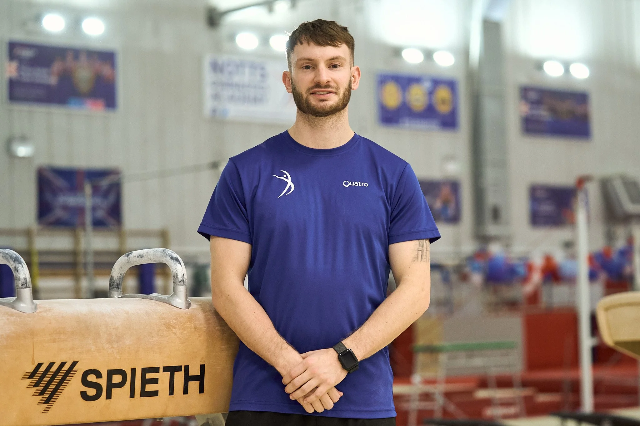 A young man with short brown hair and a beard standing in an indoor sports facility, wearing a blue athletic T-shirt with the word 'Quatro' and a logo on it, and a black watch on his left wrist. He is standing beside a wooden springboard labeled 'SPIETH'.