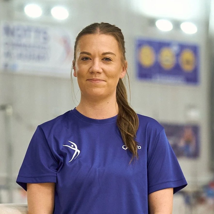 A woman with long brown hair in a ponytail, wearing a blue sports shirt, standing indoors with a blurred background of posters and lights.