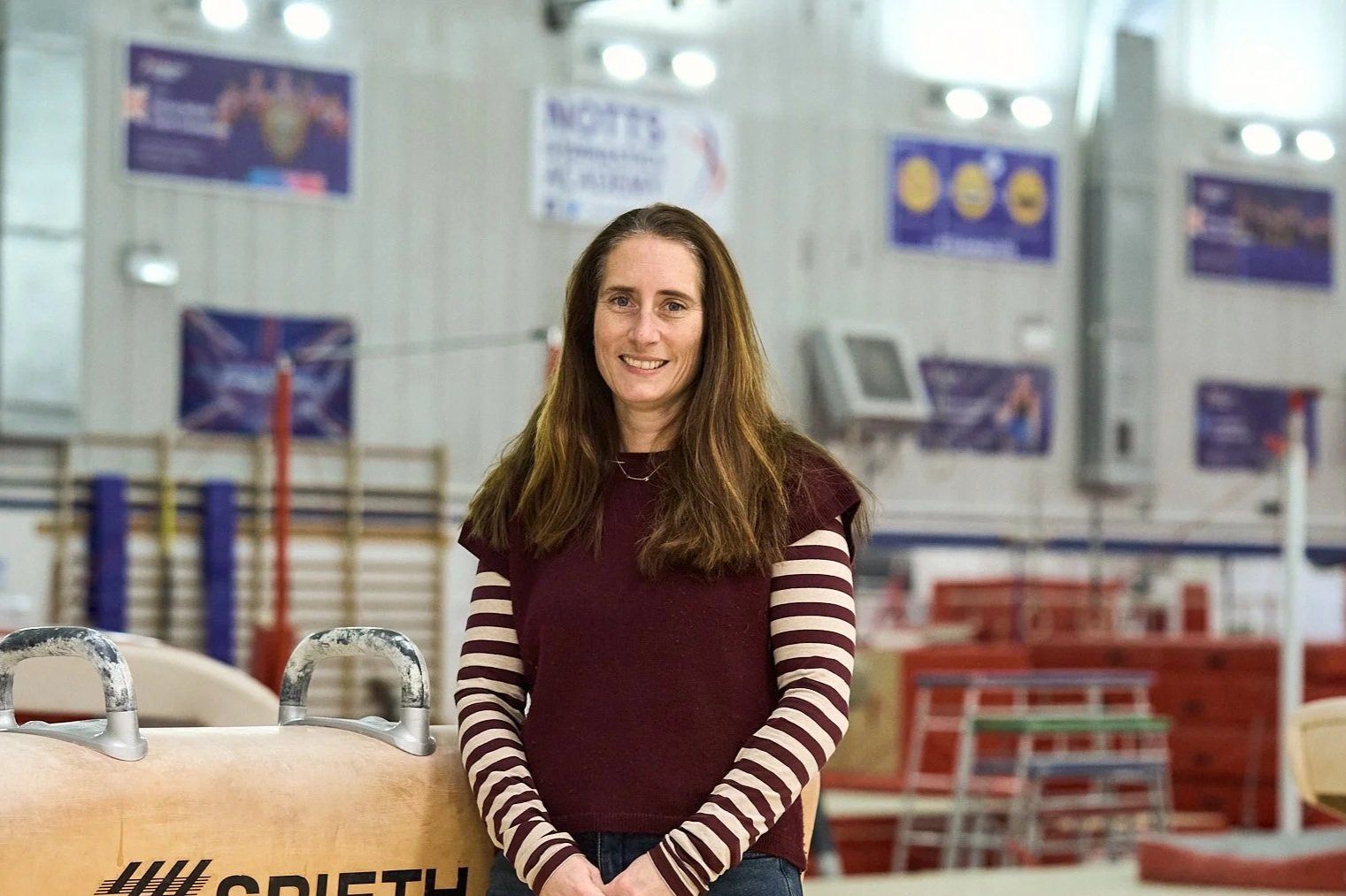 Close-up of a woman with long reddish hair, blue eyes, and freckles, wearing a dark top, against a plain light-colored background.